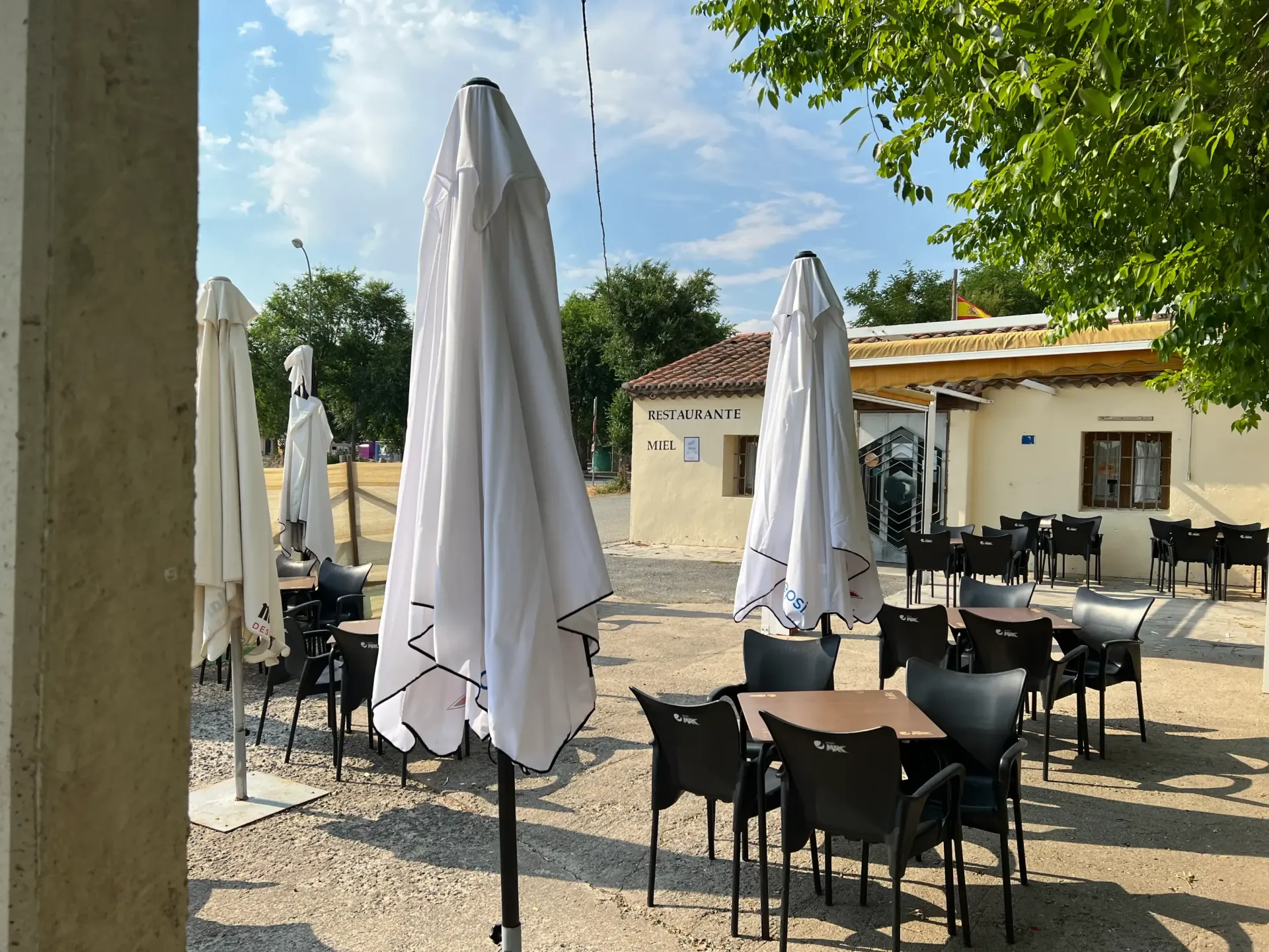 Terraza de una cafetería al aire libre con sombrillas blancas cerradas, sillas y mesas negras, bajo un cielo soleado.