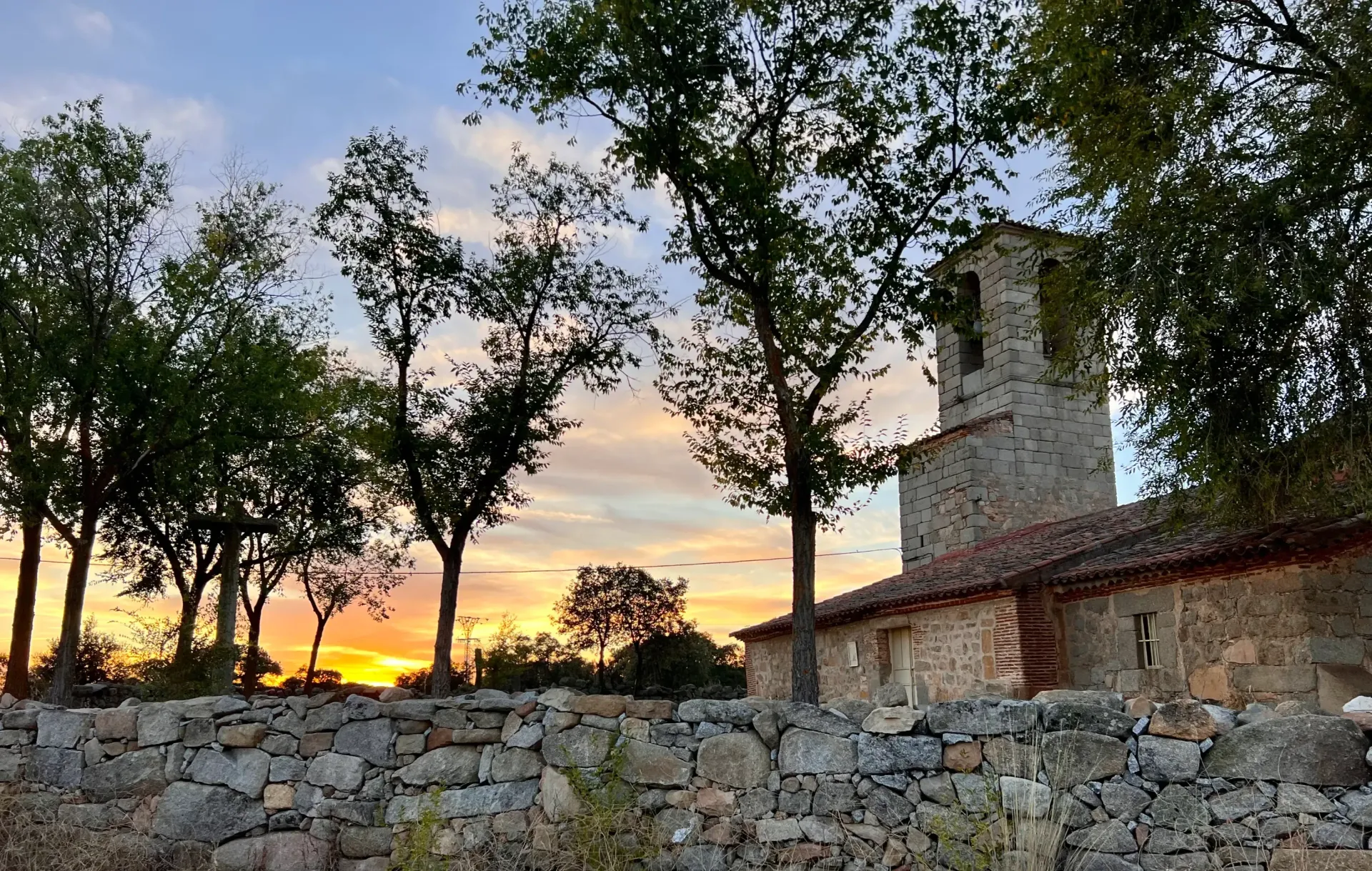 Muro de piedra y edificio rústico al atardecer, enmarcados por árboles contra un cielo colorido.