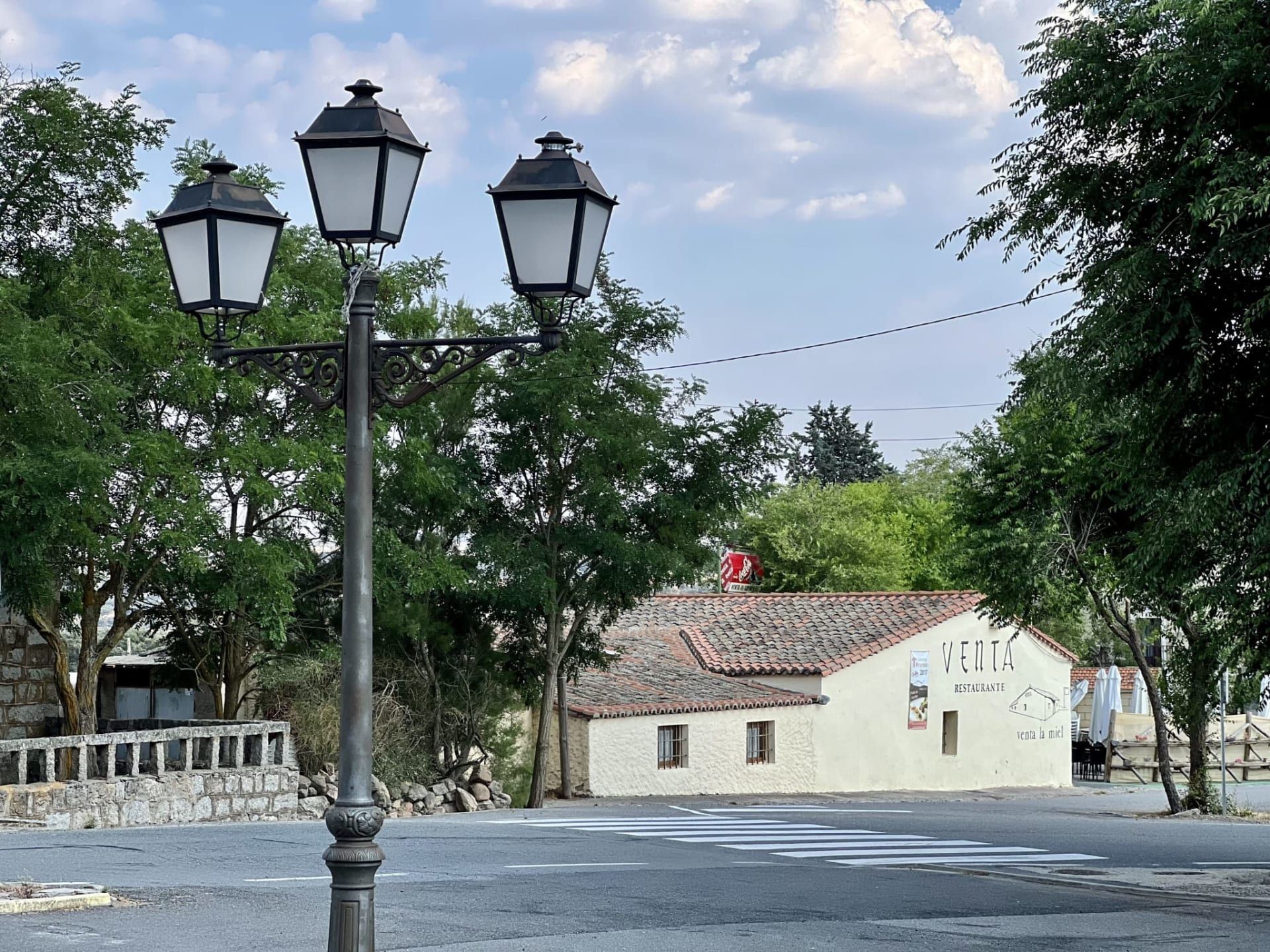 Tres farolas negras junto a una calle tranquila y un edificio beige bajo los árboles y el cielo nublado.