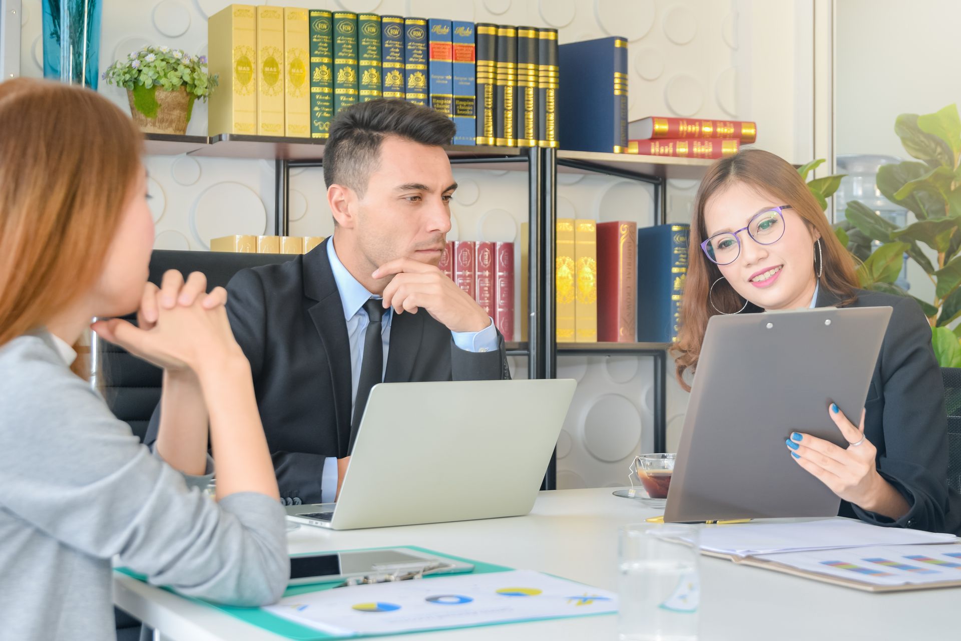 Three people in business attire at a table reviewing documents, one holding a clipboard.