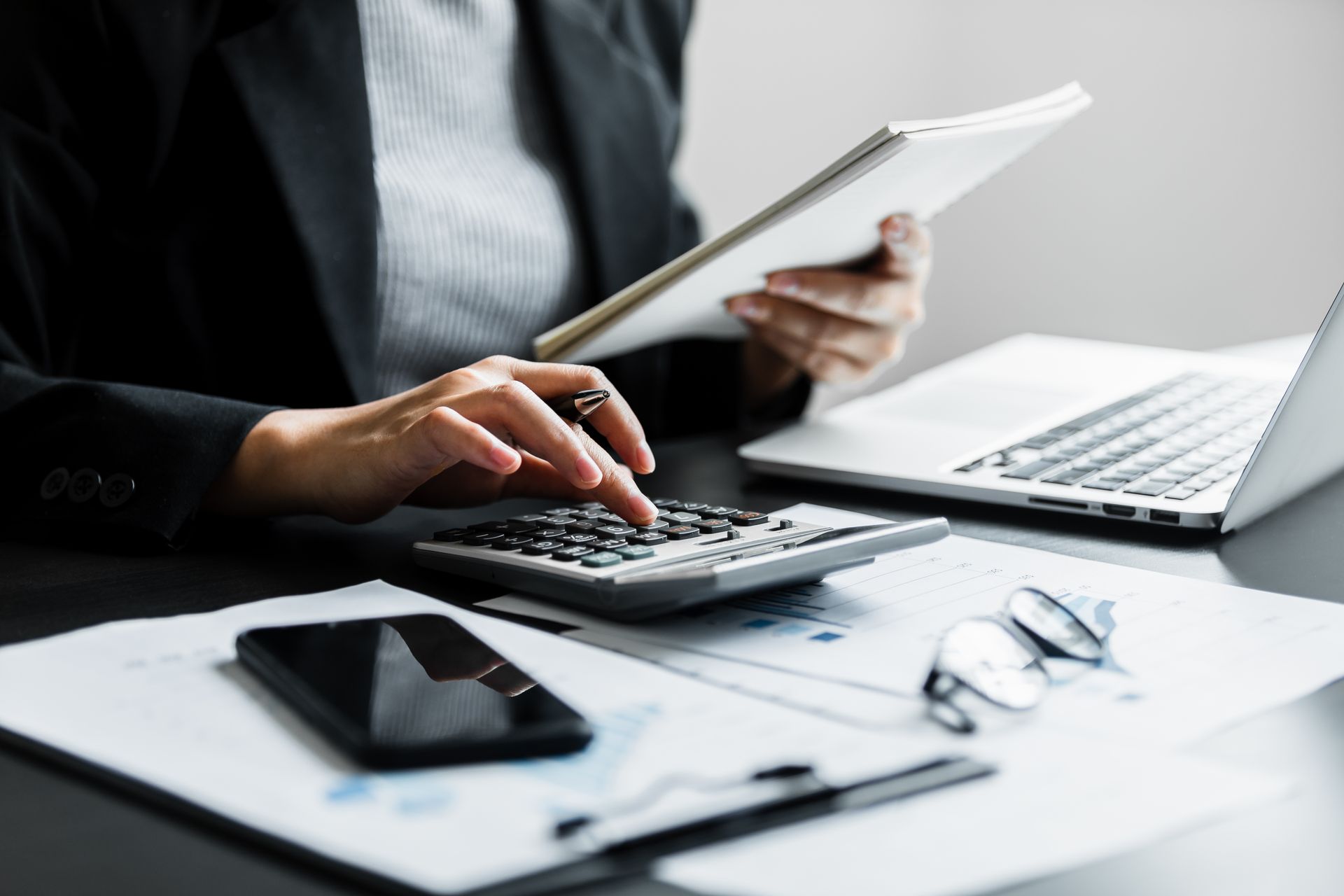 Person in a blazer using a calculator, holding a tablet, with a laptop, phone, and documents on a desk.