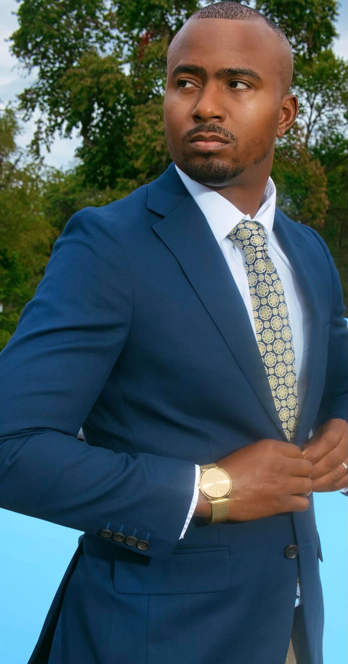 Man in blue suit, adjusting his jacket, wearing a patterned tie, gold watch, outdoors.