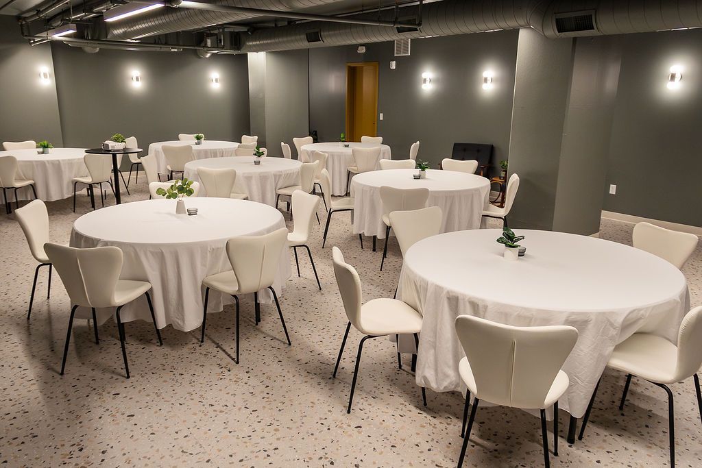 Empty banquet room with round tables covered in white cloths and white chairs under dim lighting