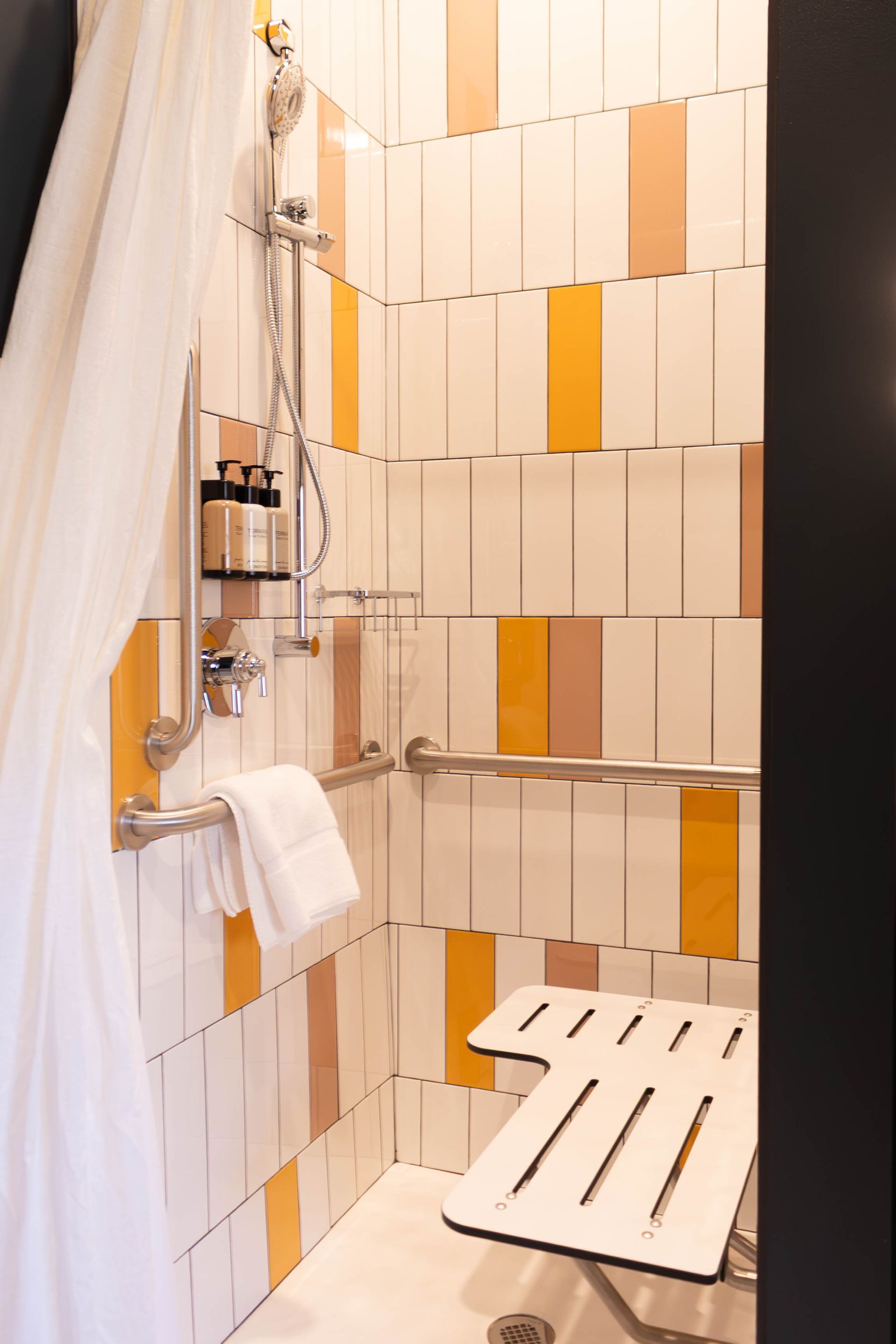 Small tiled bathroom with shower curtain, wall shelf, towels, and a wooden bath stool.