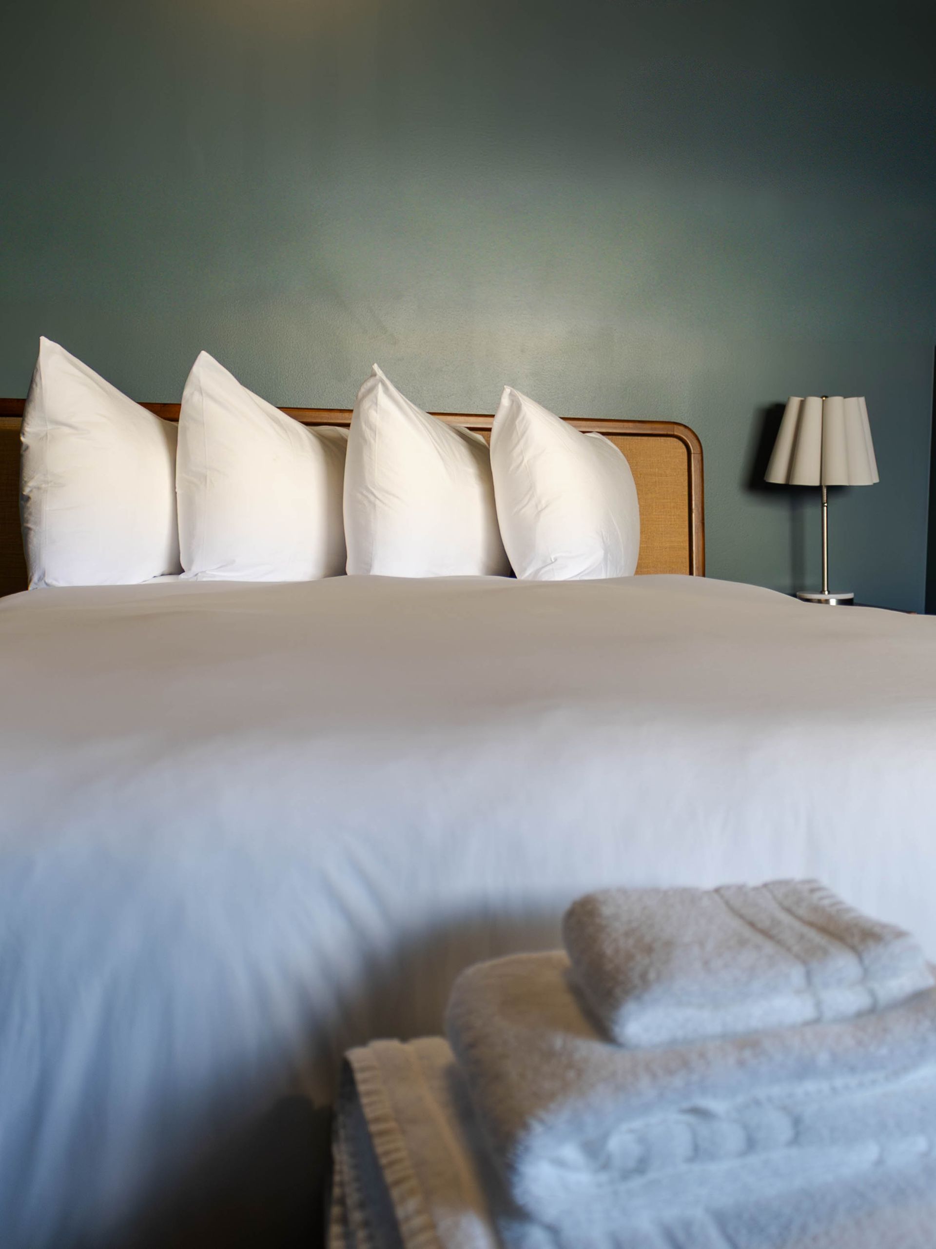 White bed with four pillows and folded towels in a softly lit bedroom