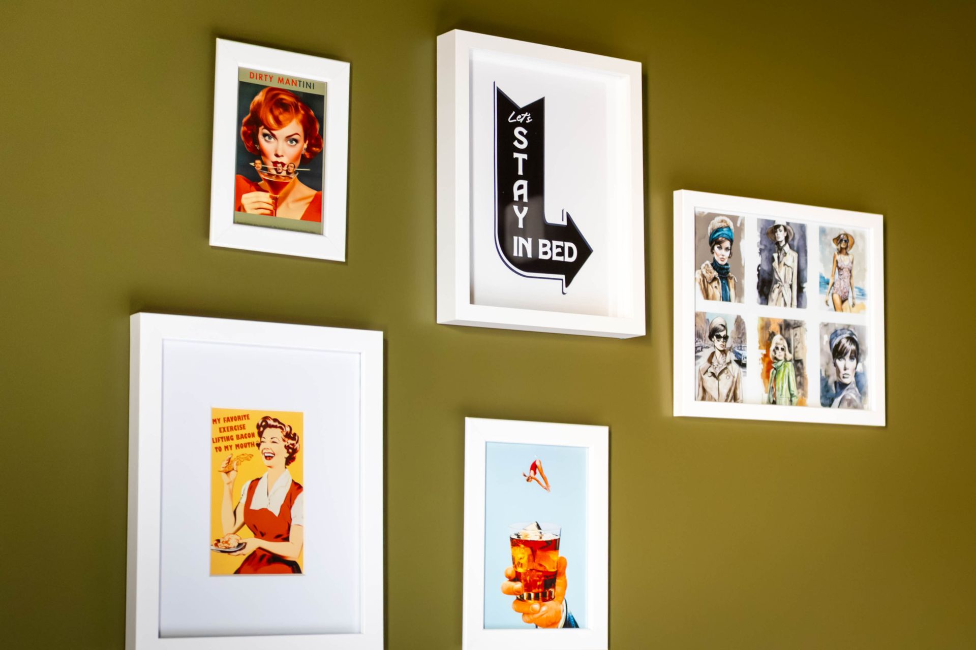 Five framed art prints arranged on an olive-green wall, including a black-and-white arrow sign and colorful illustrations