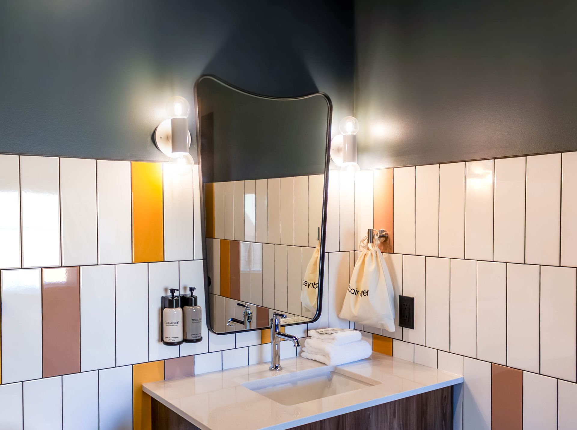 Bathroom sink with mirror, wall lights, and white-tiled backsplash with one orange accent tile