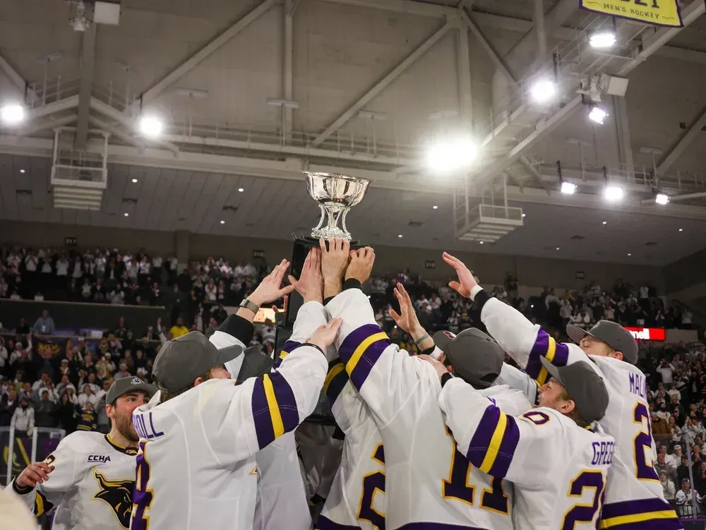 Hockey team in white and purple jerseys lifting a championship trophy on the ice