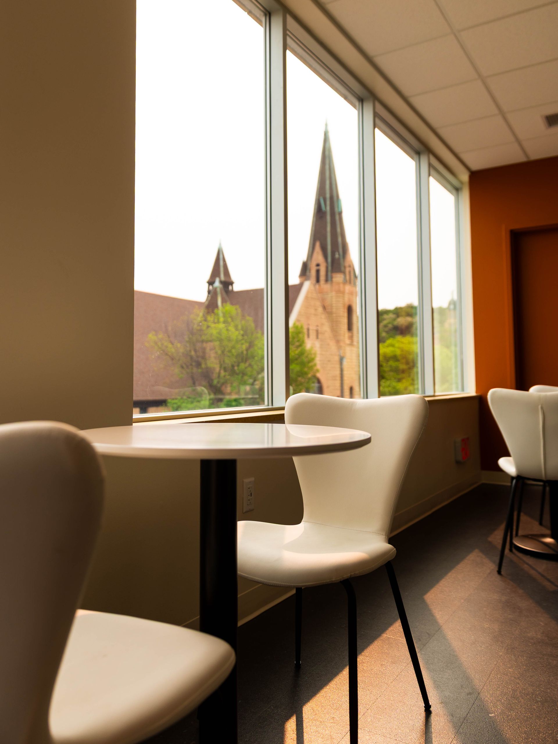 Bright café interior with white chairs and tables by large windows overlooking a church tower.