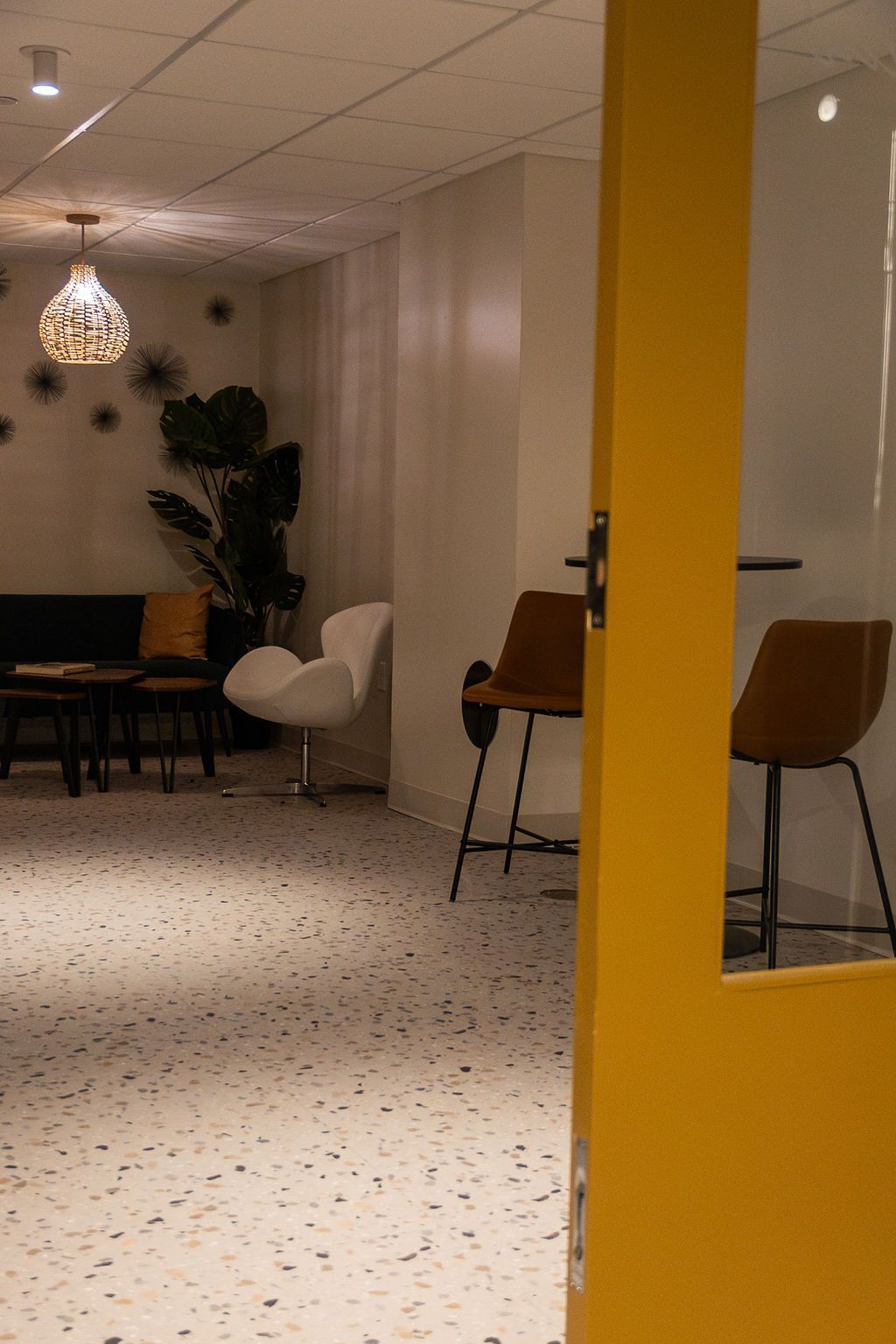 Bright hallway with yellow doorway, white chairs, wooden chairs, and a hanging lamp in a modern lounge area