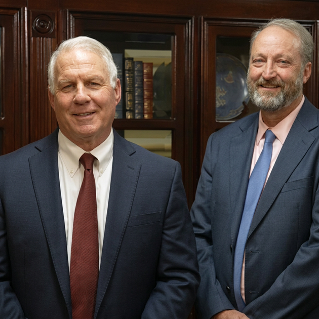 Two men in suits smiling in an office before dark wood cabinets.