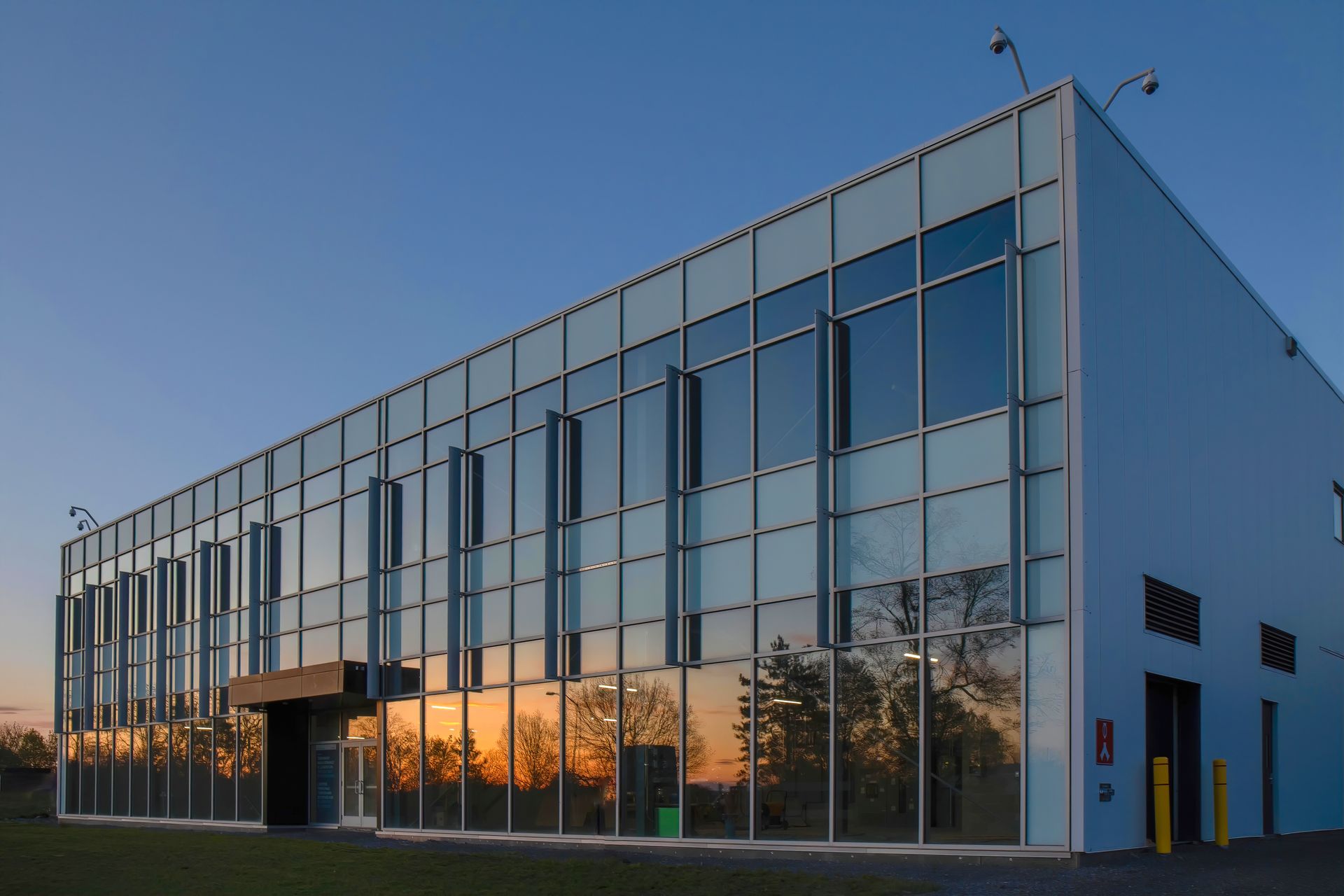 Bâtiment moderne à façade de verre, reflétant un coucher de soleil et des arbres.