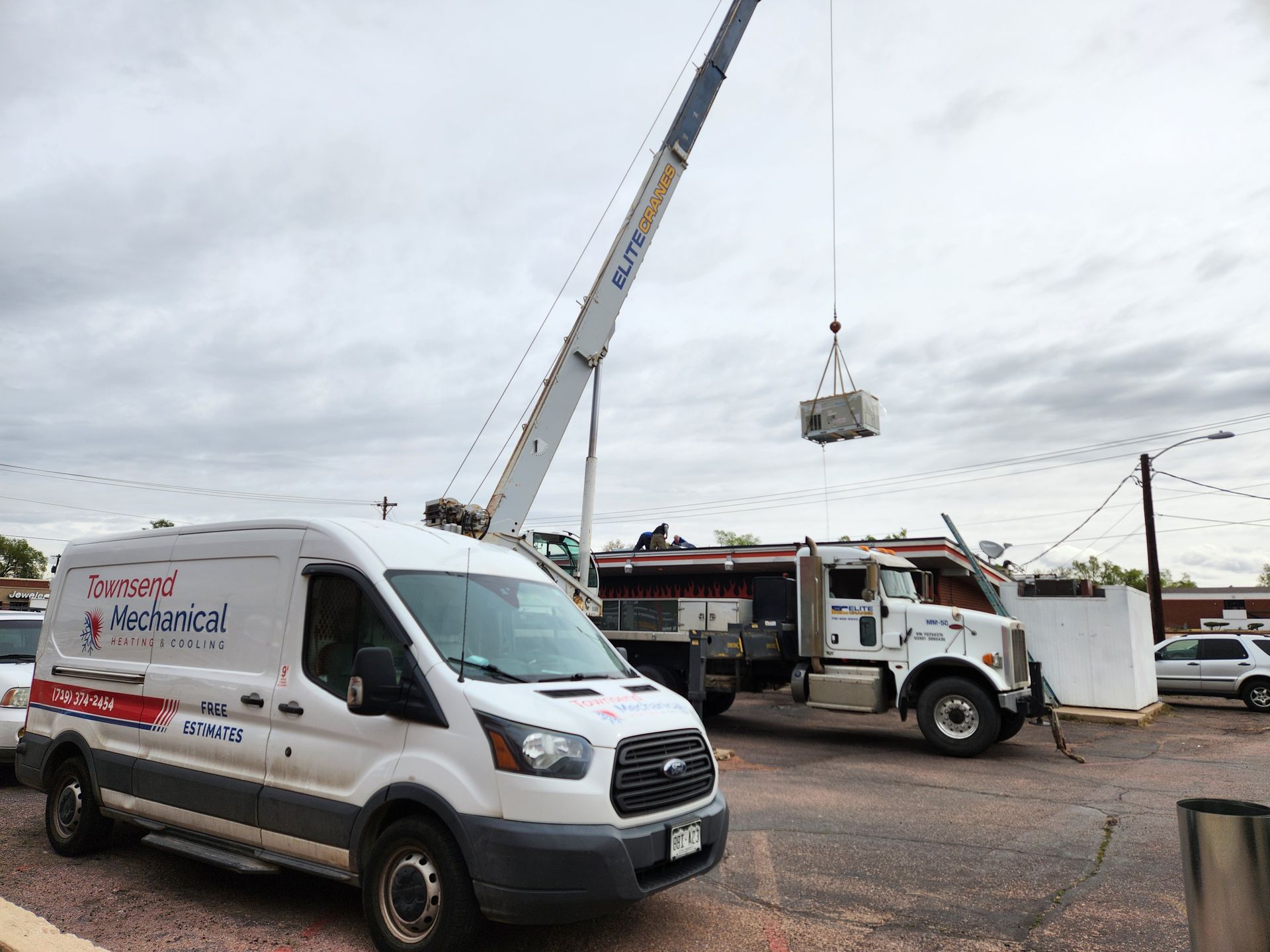 A crane lifts an HVAC unit. A service van and truck are nearby. Cloudy day.