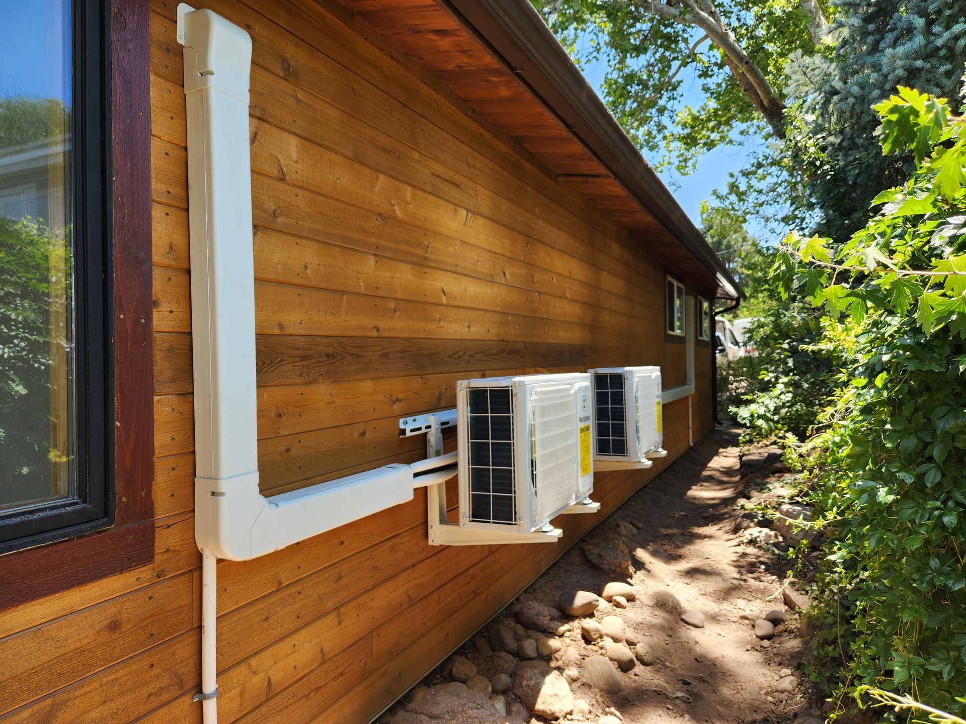Two air conditioning units mounted on a wooden building exterior, with a white conduit.
