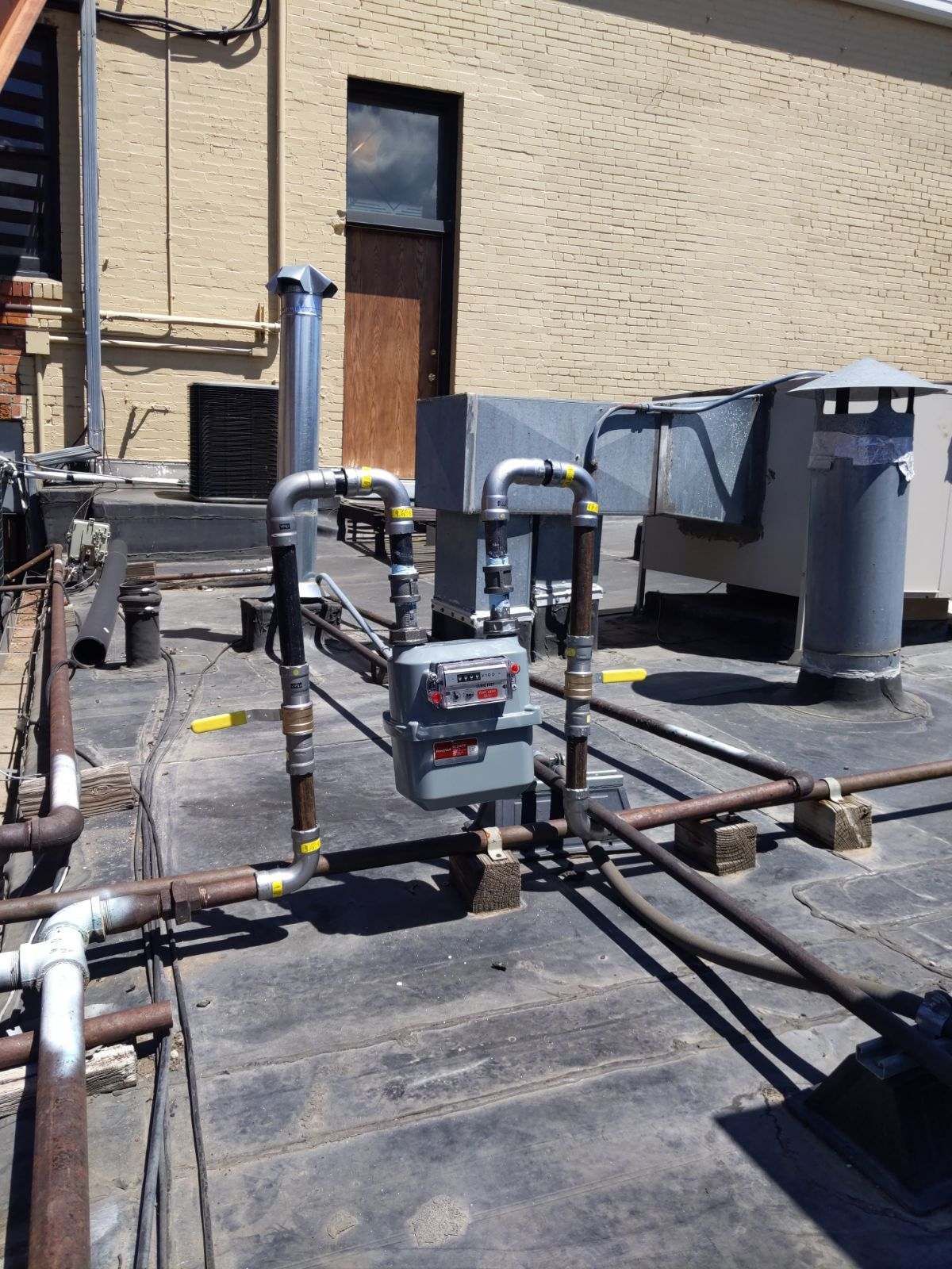 Gas meter and pipes on a building rooftop against a yellow brick wall.