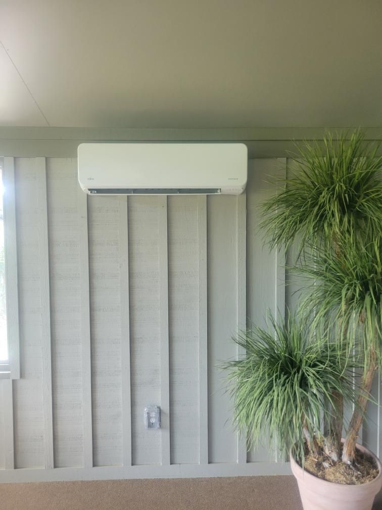 White air conditioning unit mounted on a gray vertical-paneled wall next to a potted plant.