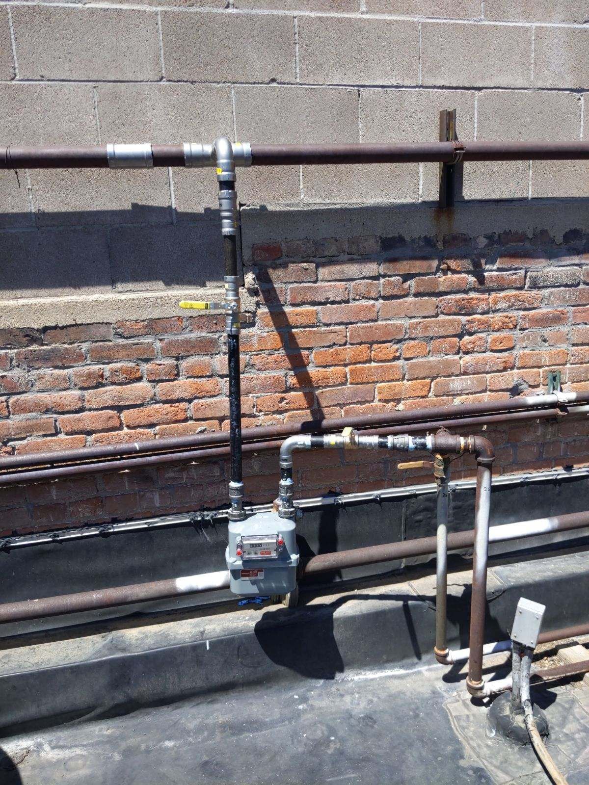 Gas meter and pipes mounted on a brick wall, attached to a dark, flat surface.
