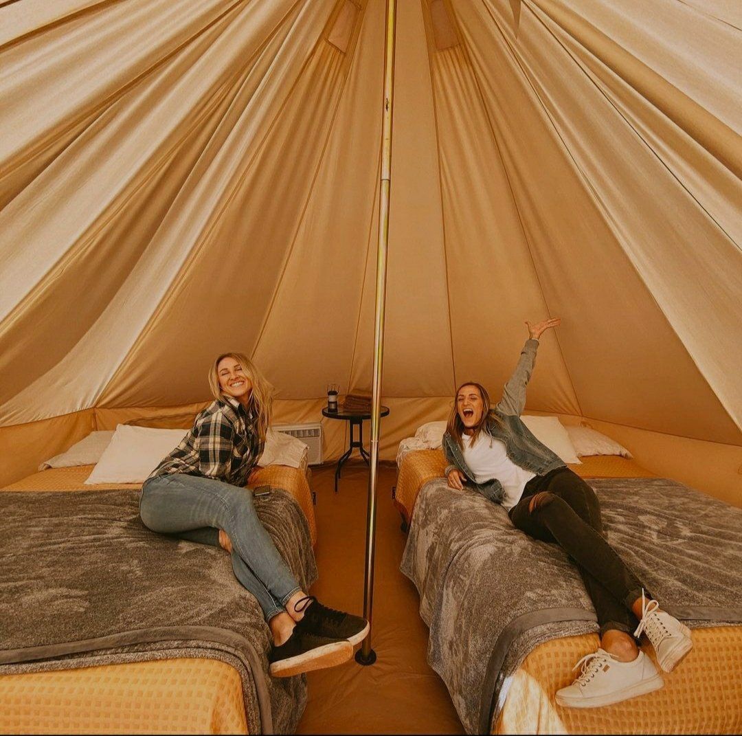 Two excited guests sitting on separate beds inside a spacious glamping tent, with big smiles!