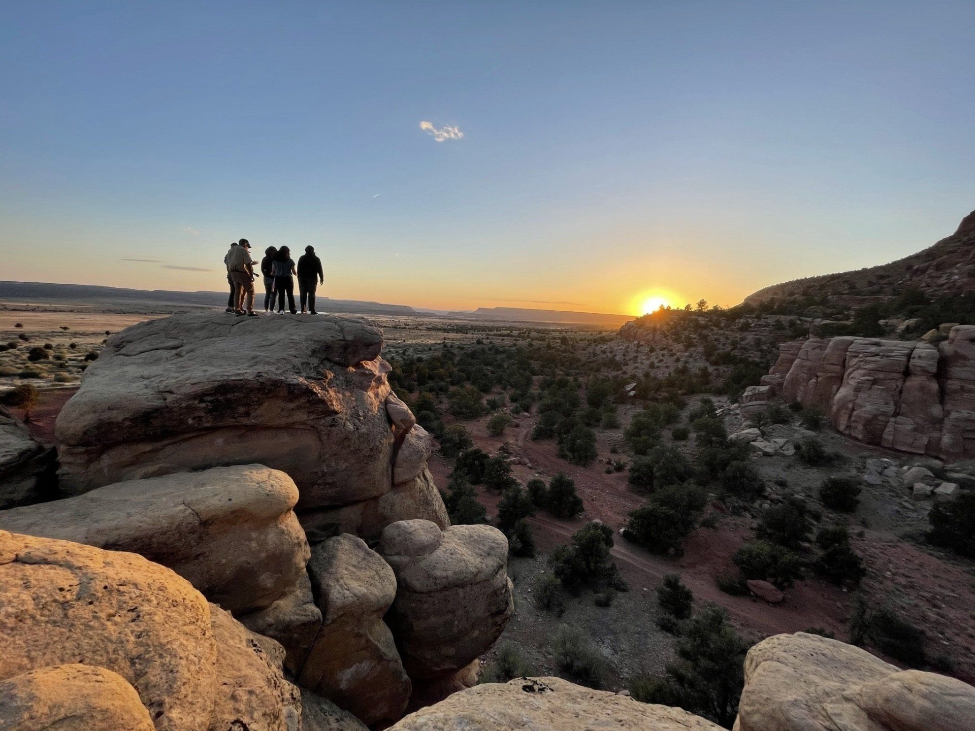 A group of people standing on a large rock formation, enjoying a sunset view over a vast landscape.