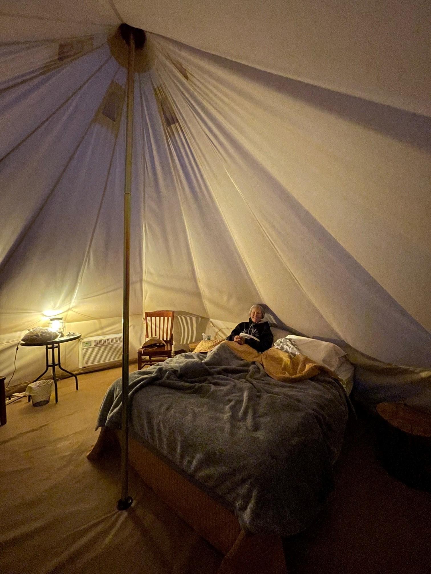 A guest relaxing on a bed inside a glamping tent at night, with warm lighting creating a cozy and tranquil atmosphere.