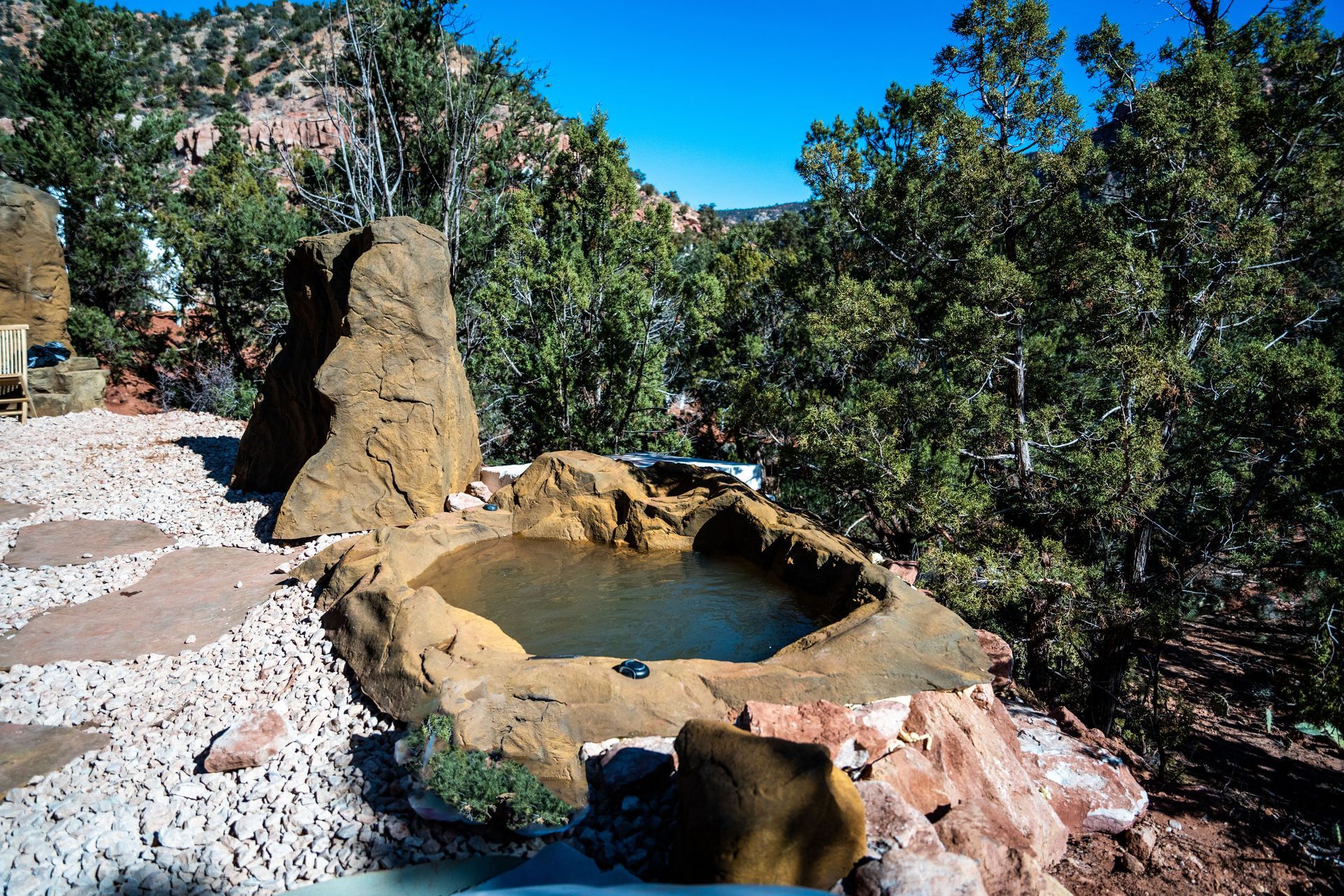 A natural rock hot tub surrounded by trees and mountains under a clear blue sky near Zion Glamping Adventures.