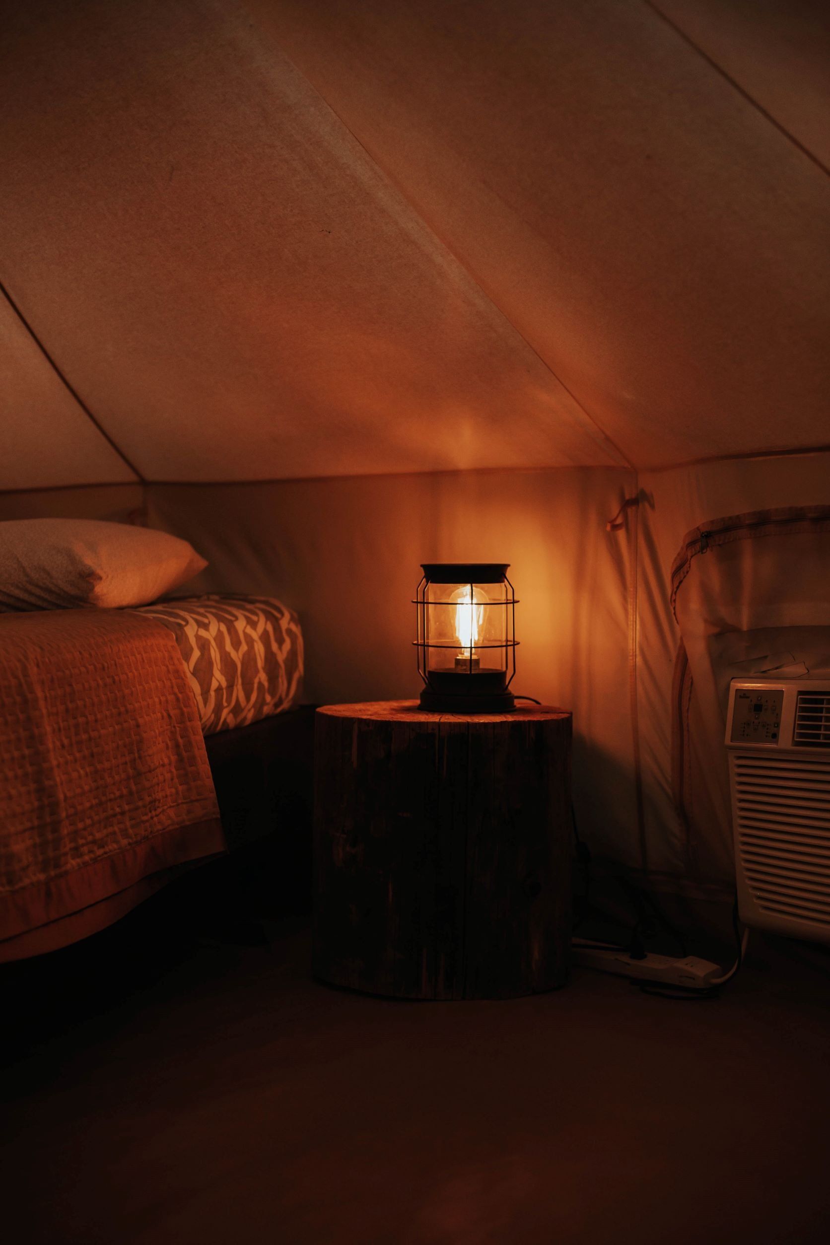 Warmly lit interior of a glamping tent, featuring a comfortable bed, a rustic lantern on a wooden nightstand.