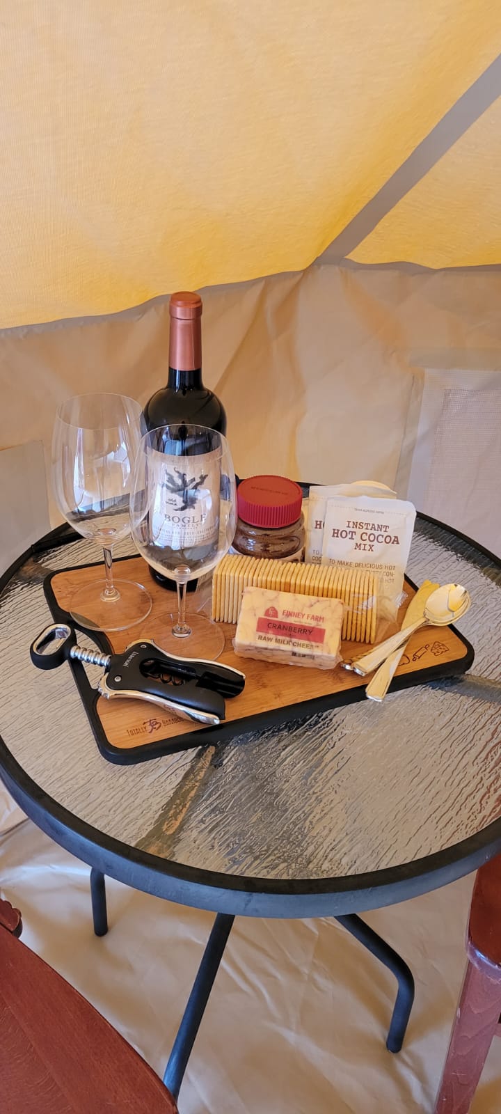 Close-up of a table set with wine, glasses, crackers, cheese, and hot cocoa mix in a glamping tent. Zion Glamping Adventures.