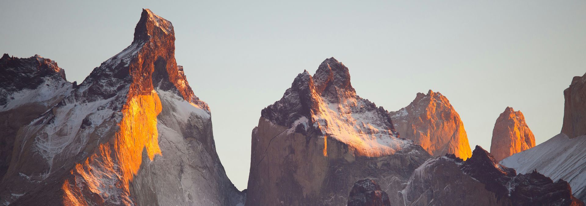Snowy mountain peaks lit by orange sunset glow under a pale sky