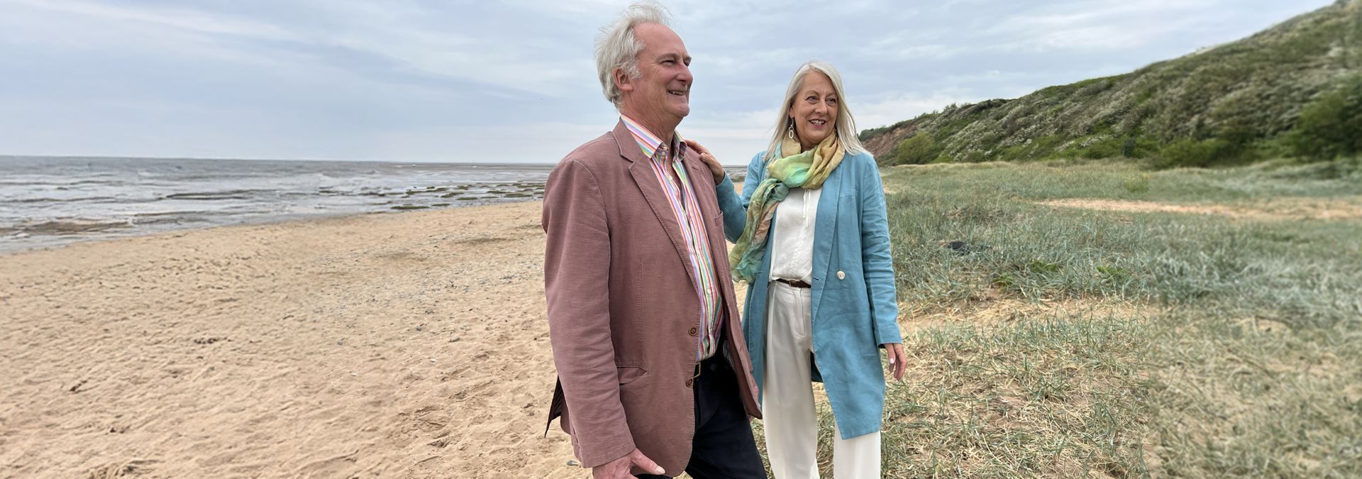 Two people standing on a sandy beach beside grassy dunes under an overcast sky