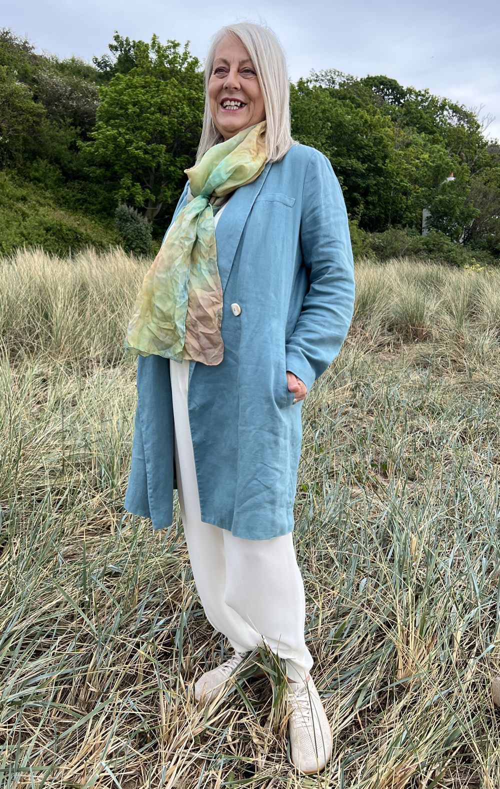 Woman standing outdoors in a light blue coat and scarf, smiling in a grassy field.