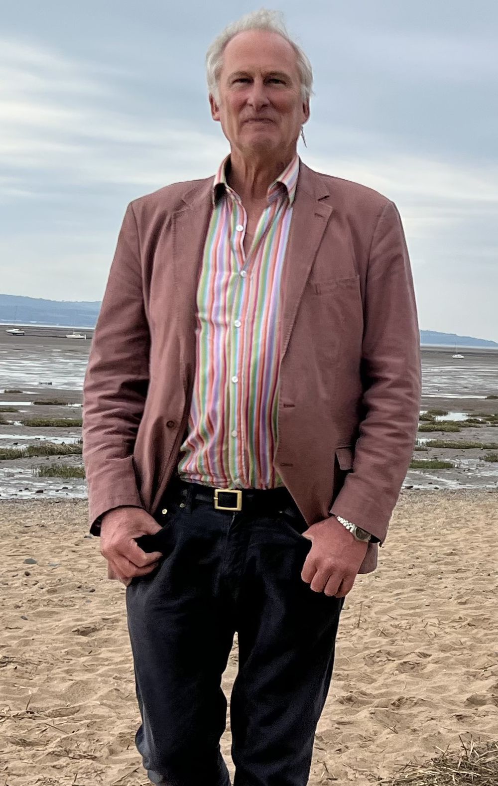 Man standing on a beach in a pink jacket and striped shirt, with sea and cloudy sky behind him