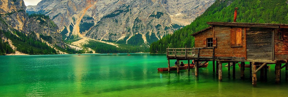Wooden cabin on stilts beside a vivid green lake with mountains in the background