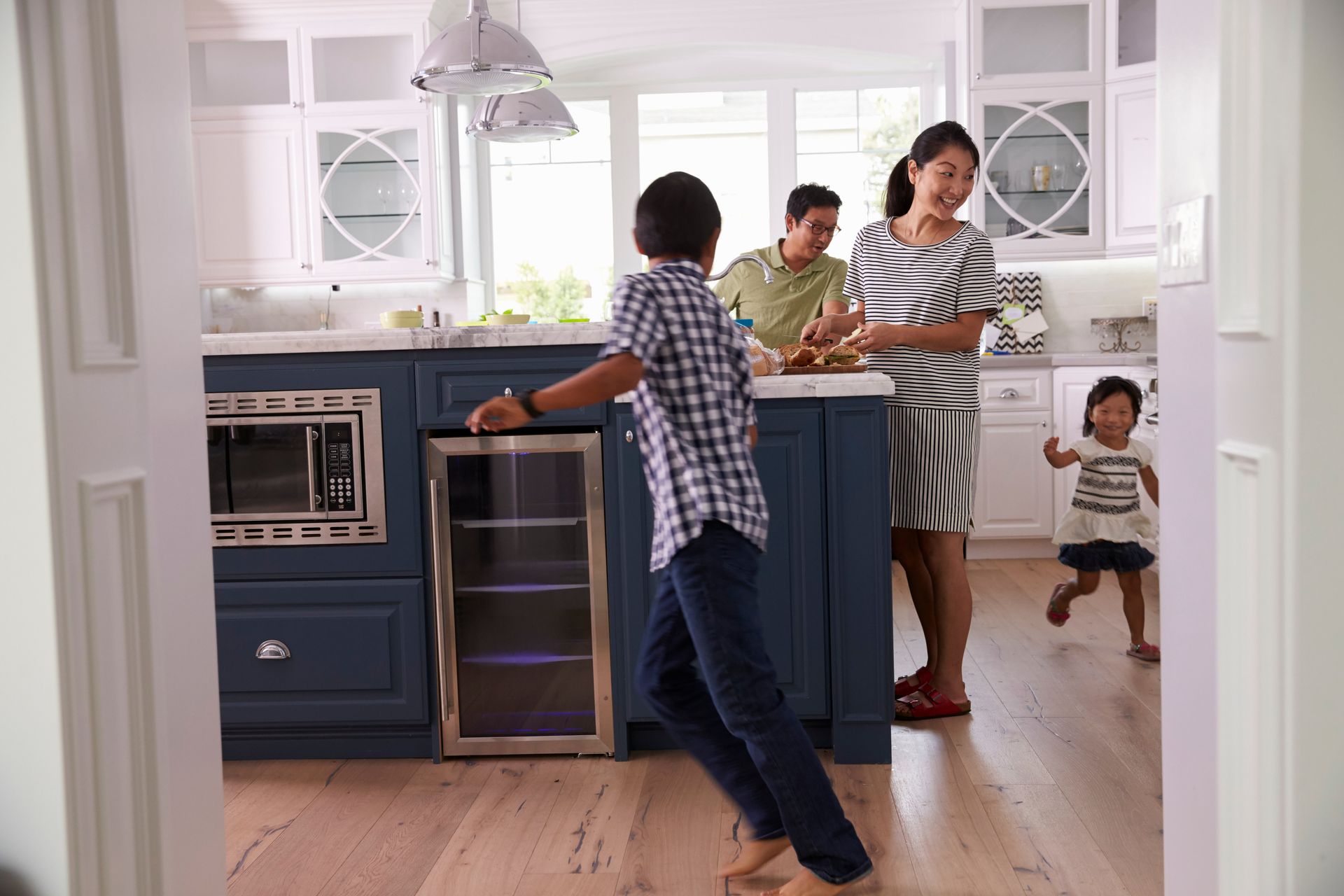 A small mouse sits inside a wire cage trap set on a grassy outdoor surface. A family moves through a bright kitchen featuring blue cabinets, a built-in microwave, and a glass-front beverage cooler.