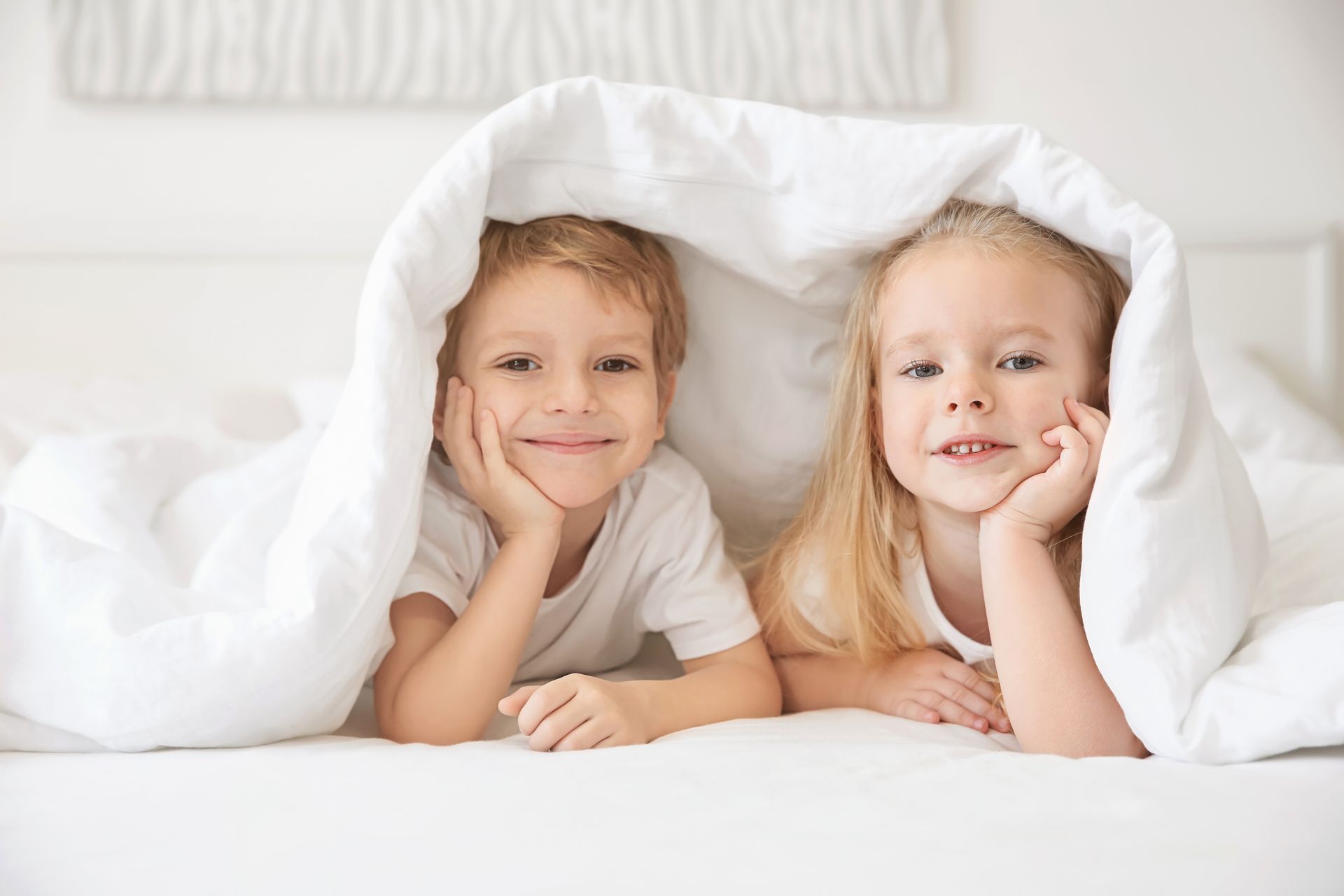 Two children with smiling expressions resting their chins on their hands while nestled together under a white blanket.