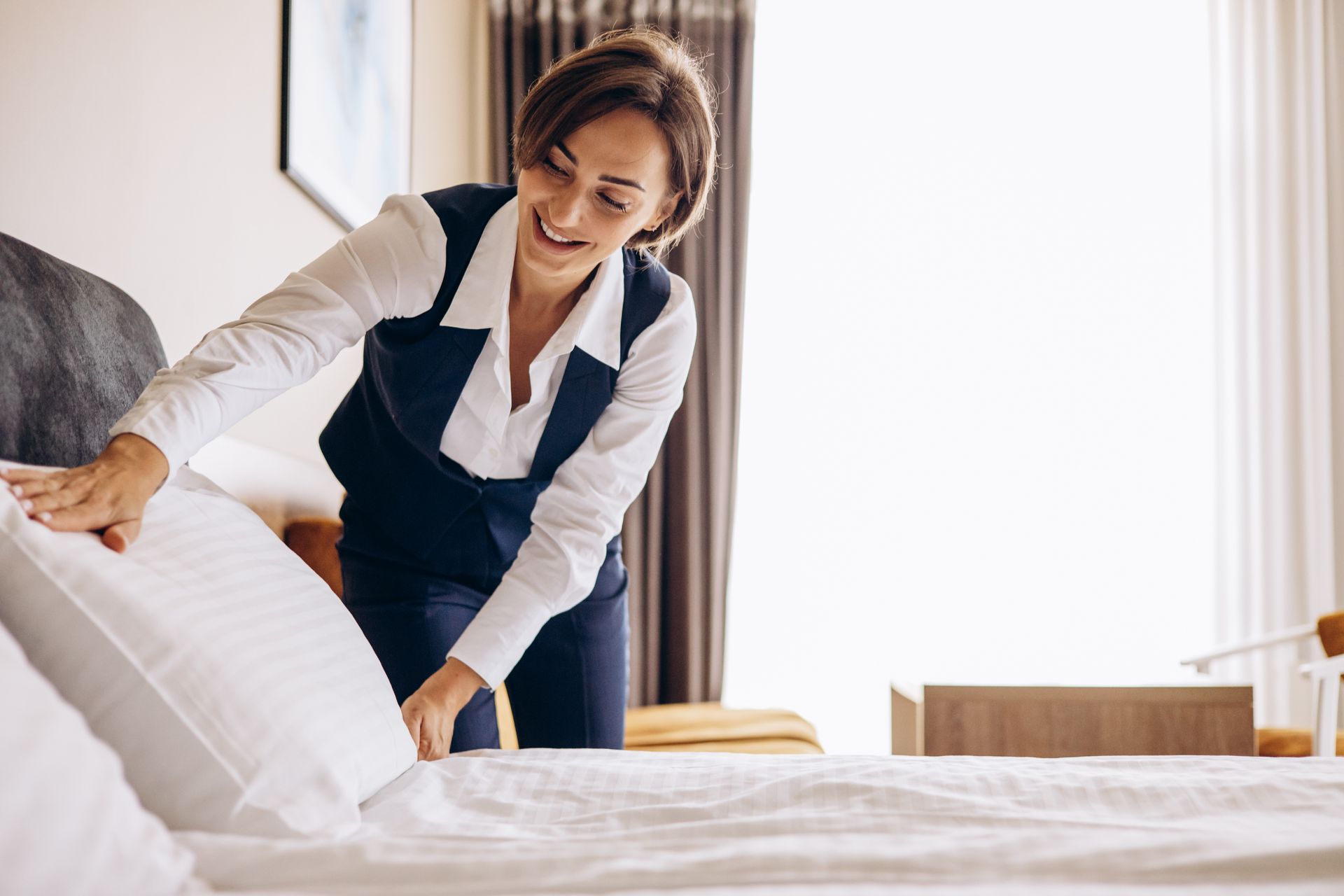 A hotel housekeeper in a white shirt and dark vest smiling while arranging a pillow on a neatly made bed in a guest room.