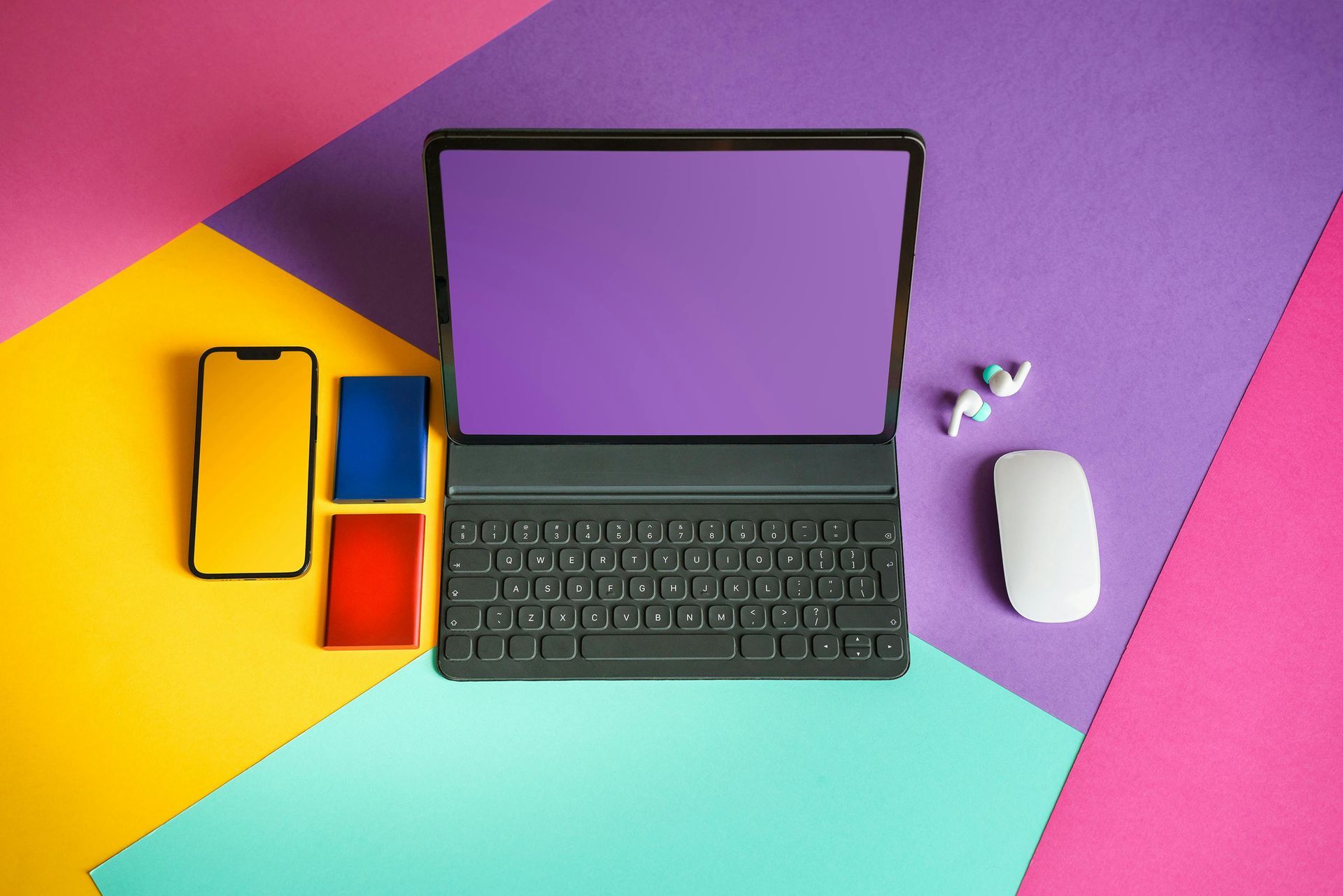 A laptop , phone , mouse and ear buds on a colorful background.