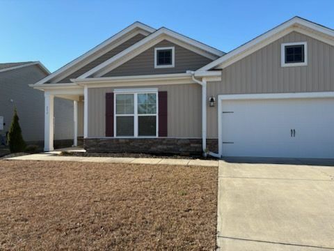 A single-story suburban house with beige vertical siding, stone accents, a white garage door, and a brown lawn.