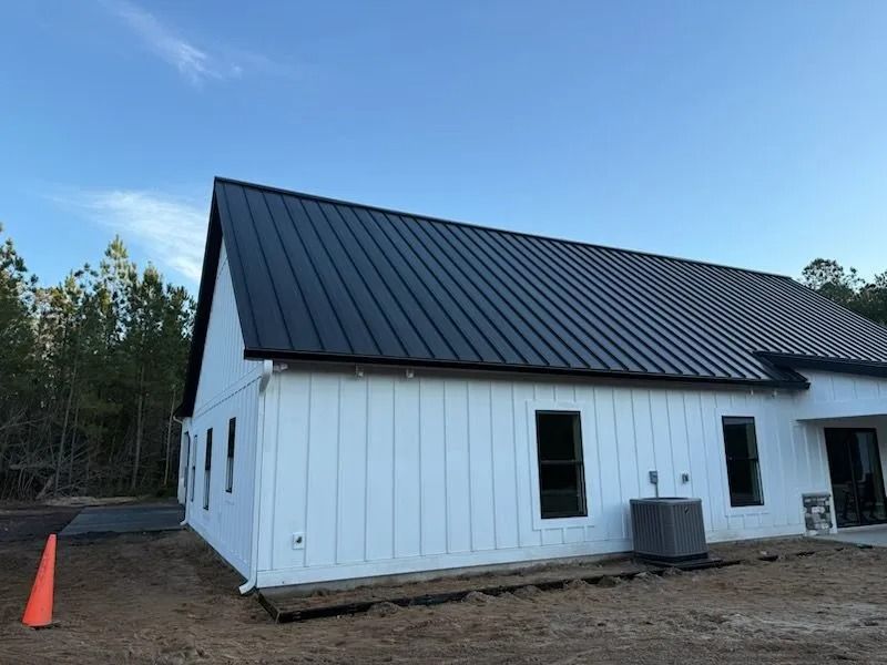 A white farmhouse-style home with a black metal roof under a clear blue sky, set on a construction lot.