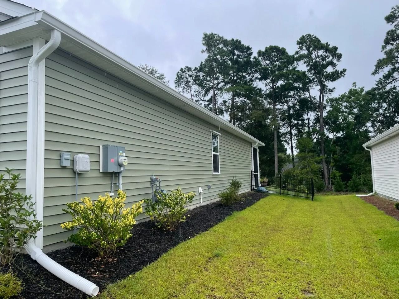The side of a sage-green house with white trim, a downspout, and a landscaped mulch bed bordering a grassy lawn.