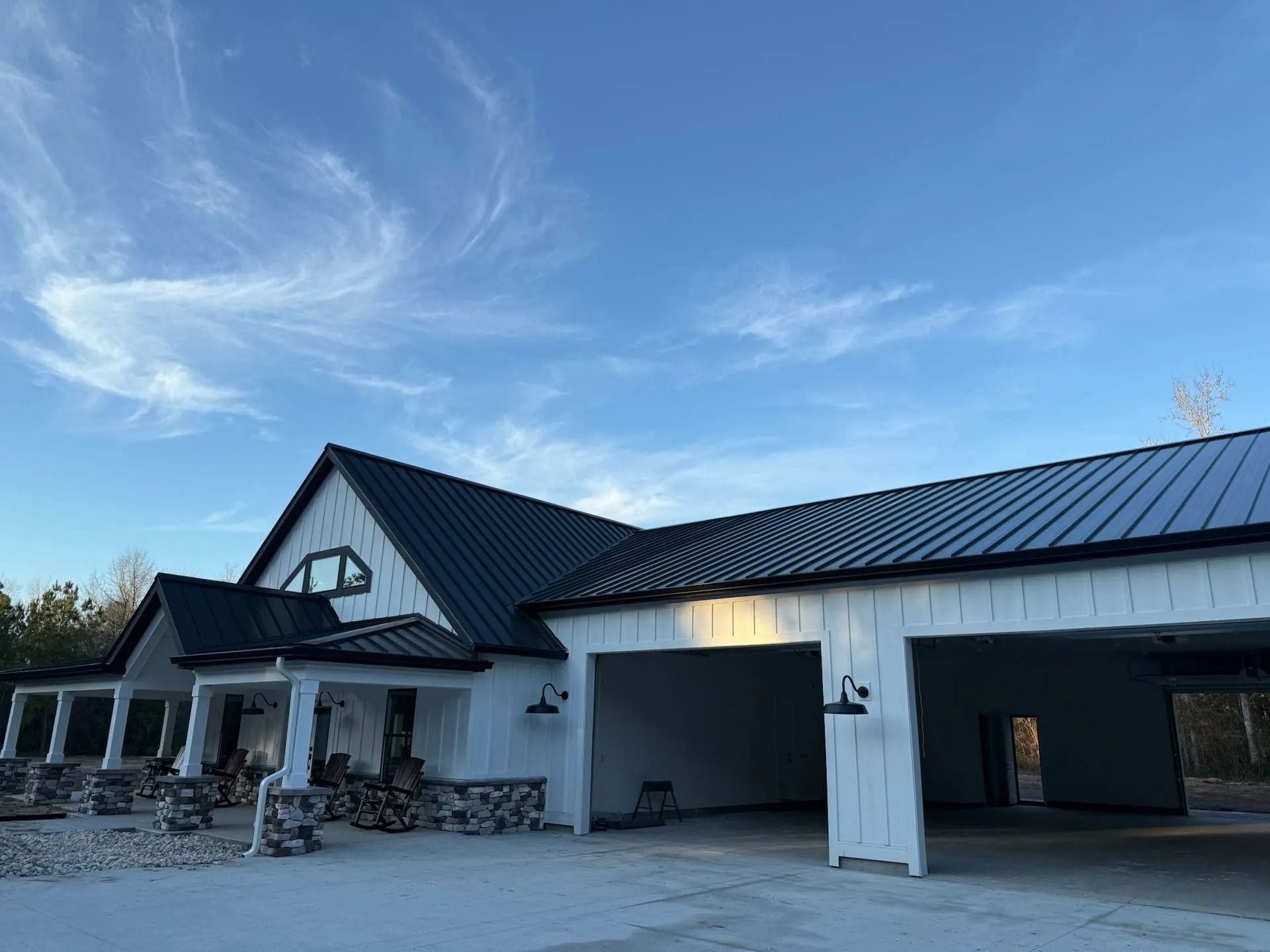 White farmhouse with a black metal roof, a wrap-around porch, and an attached multi-car garage under a clear blue sky.