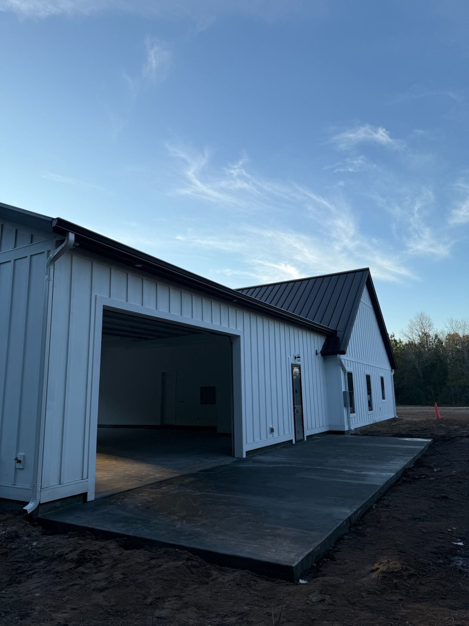 A white pole-barn style building with a black roof and a freshly poured concrete patio under a blue, sunny sky.