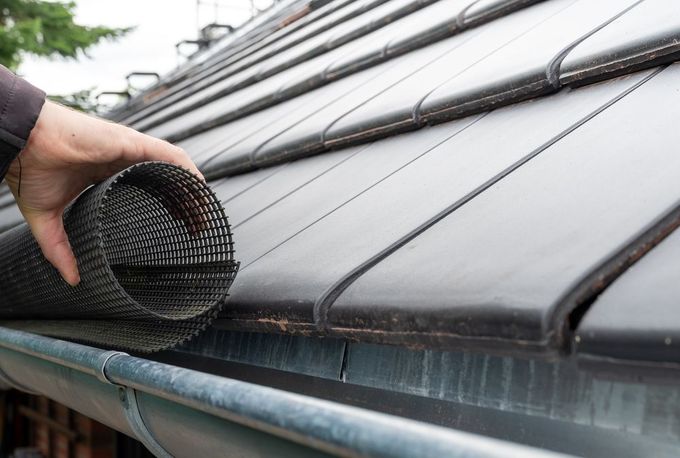 A hand installing black plastic gutter guard mesh along the edge of a tiled roof.