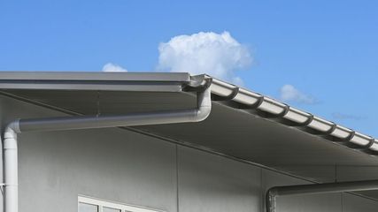 A grey metal gutter system attached to the roofline of a building against a bright blue sky with a white cloud.