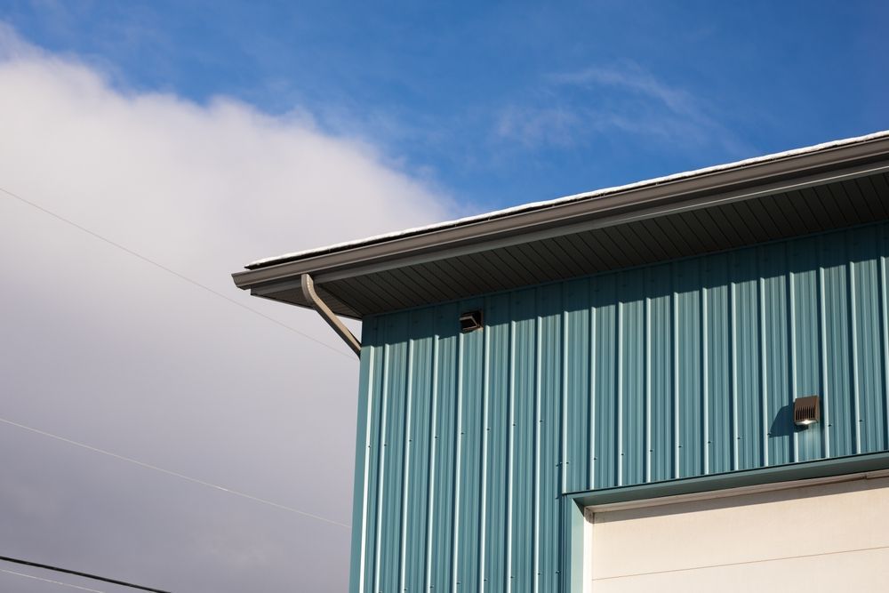The corner of a blue, corrugated metal building with a white garage door against a bright blue sky with a white cloud.