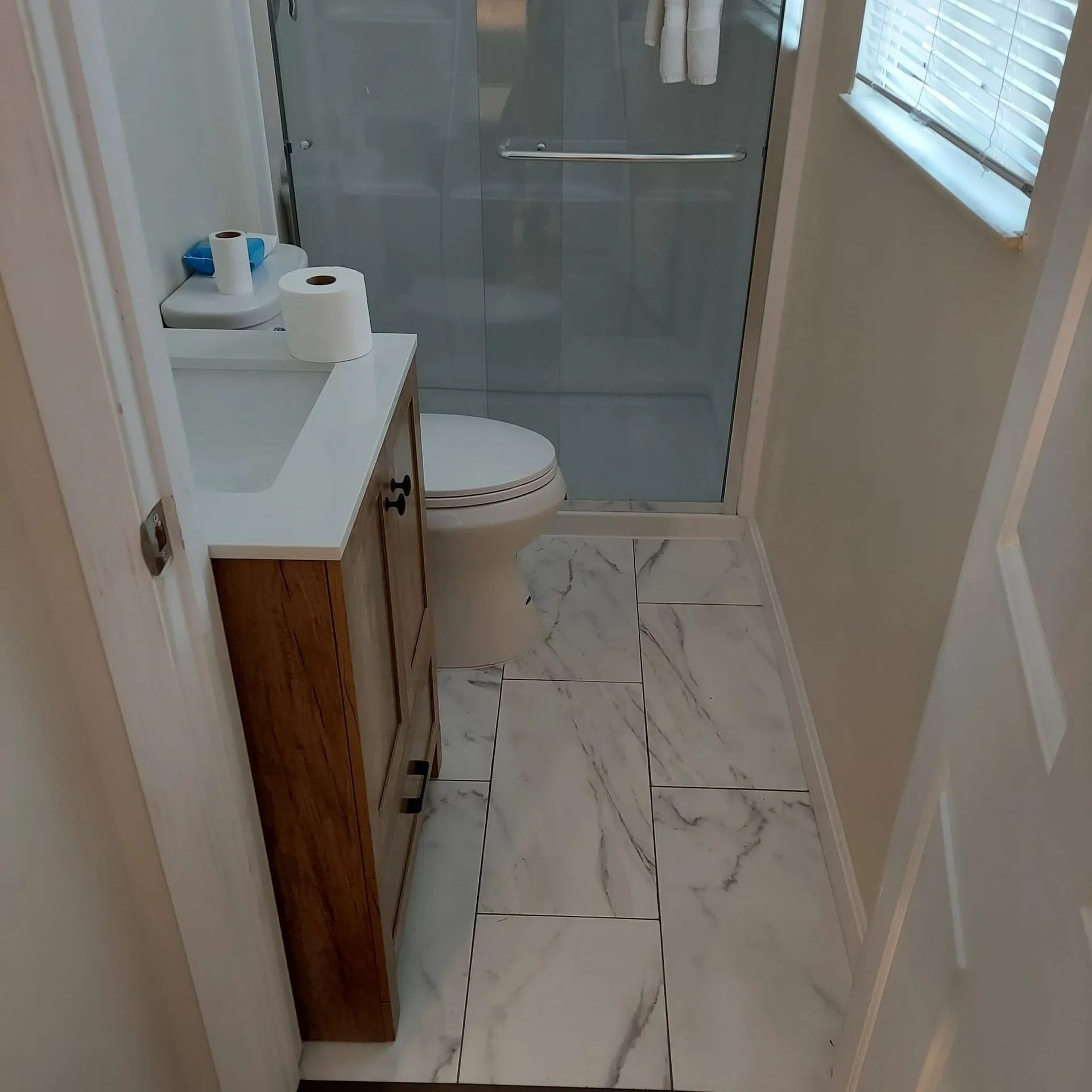 Small bathroom with glass shower, white vanity, and marble-pattern tile floor.