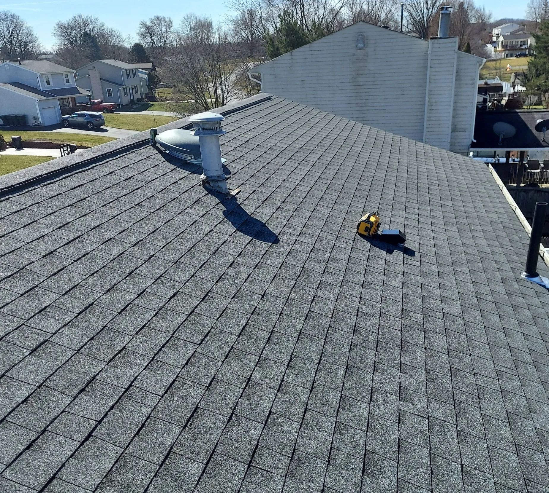 Roof with gray shingles, a worker in white on the left, and a yellow tool on the right under a clear sky