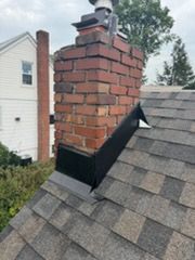 Red brick chimney with black flashing on a gray shingled roof near a white house.