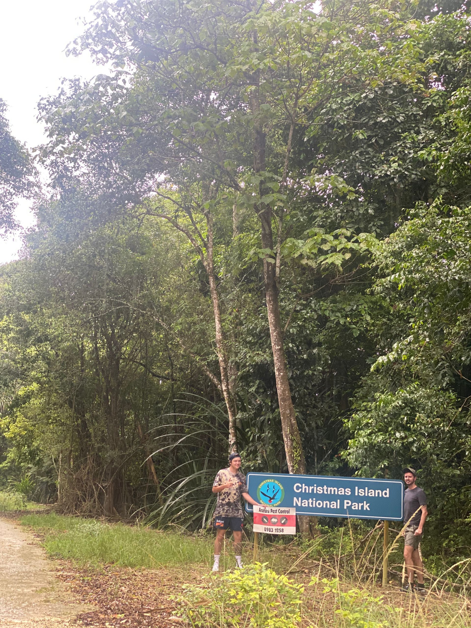 A couple of people standing next to a sign in the middle of a forest.