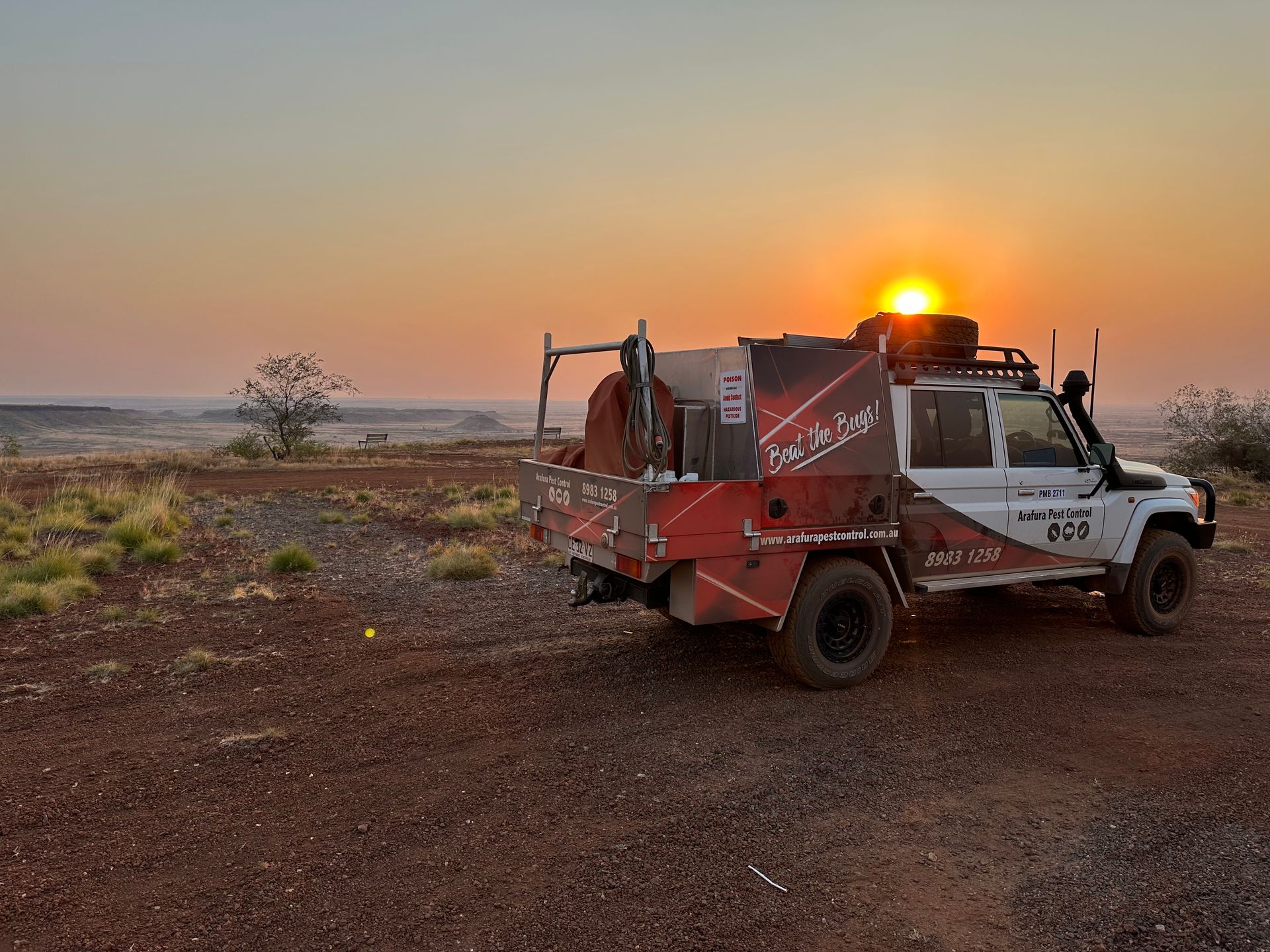 A truck is parked in a dirt field with the sun setting in the background.