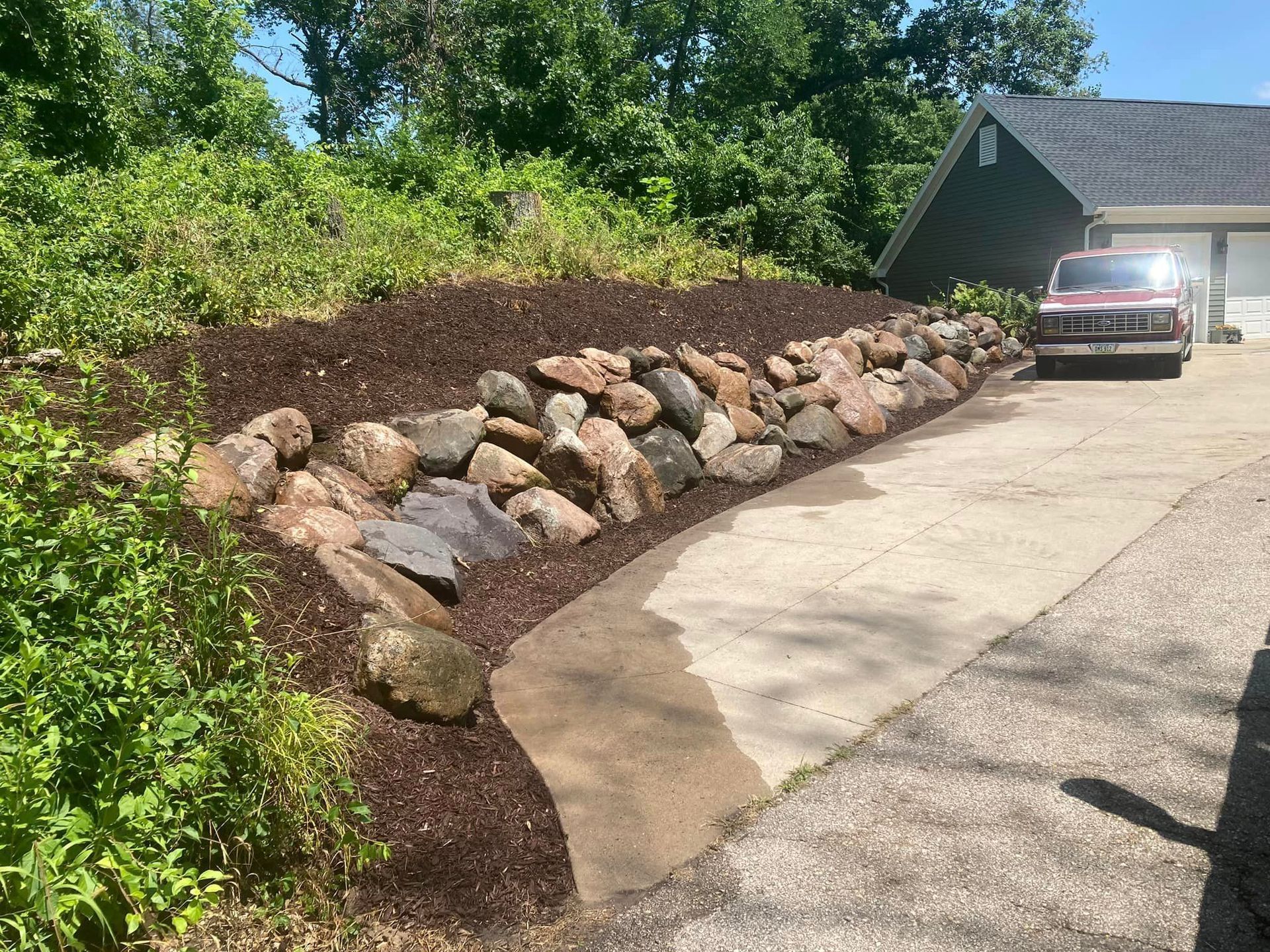 Rock retaining wall with driveway, mulch, and landscaping. A vehicle is parked near a house.